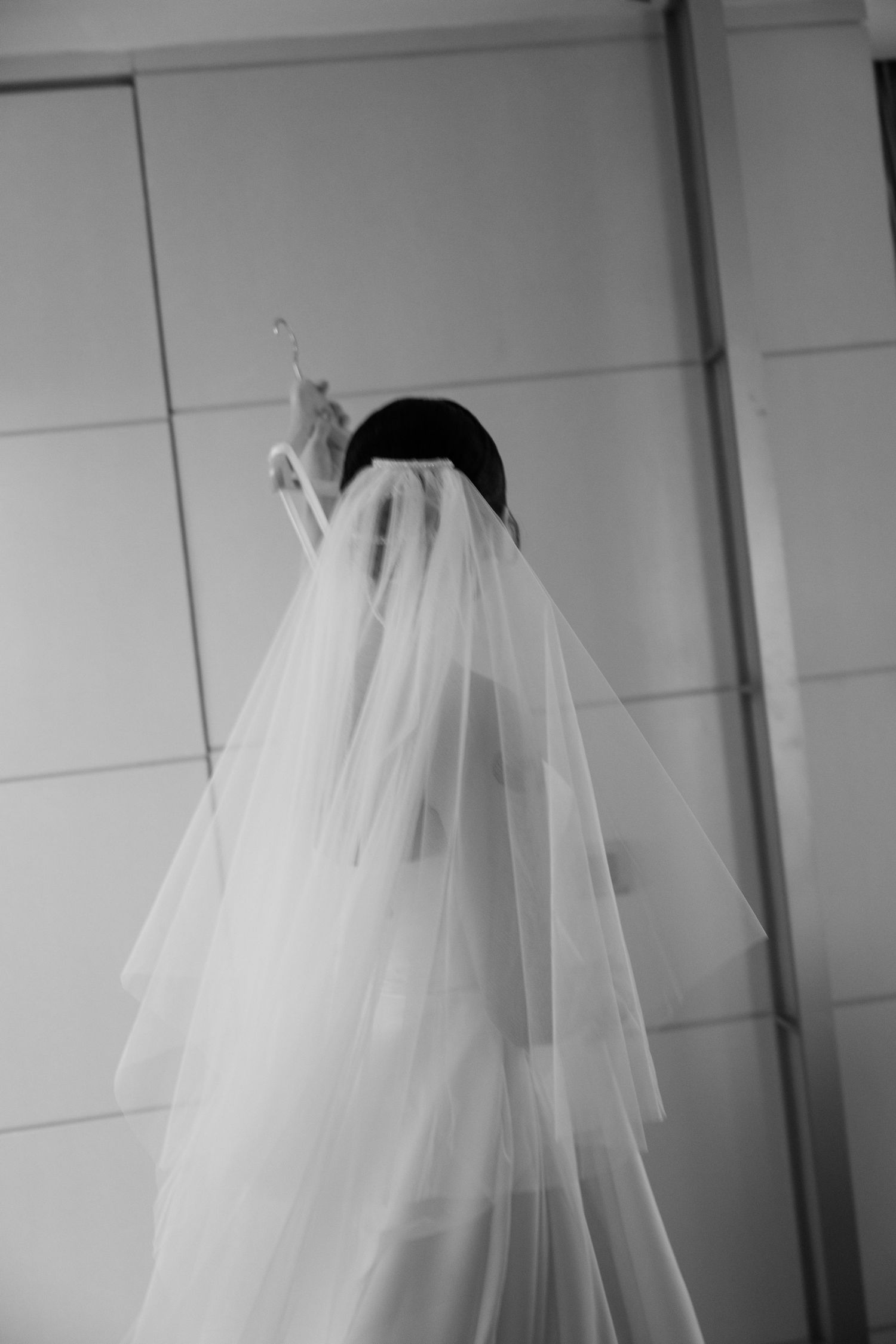 Black and white photo of a bride wearing a flowing layered wedding veil, seen from behind reaching upward.