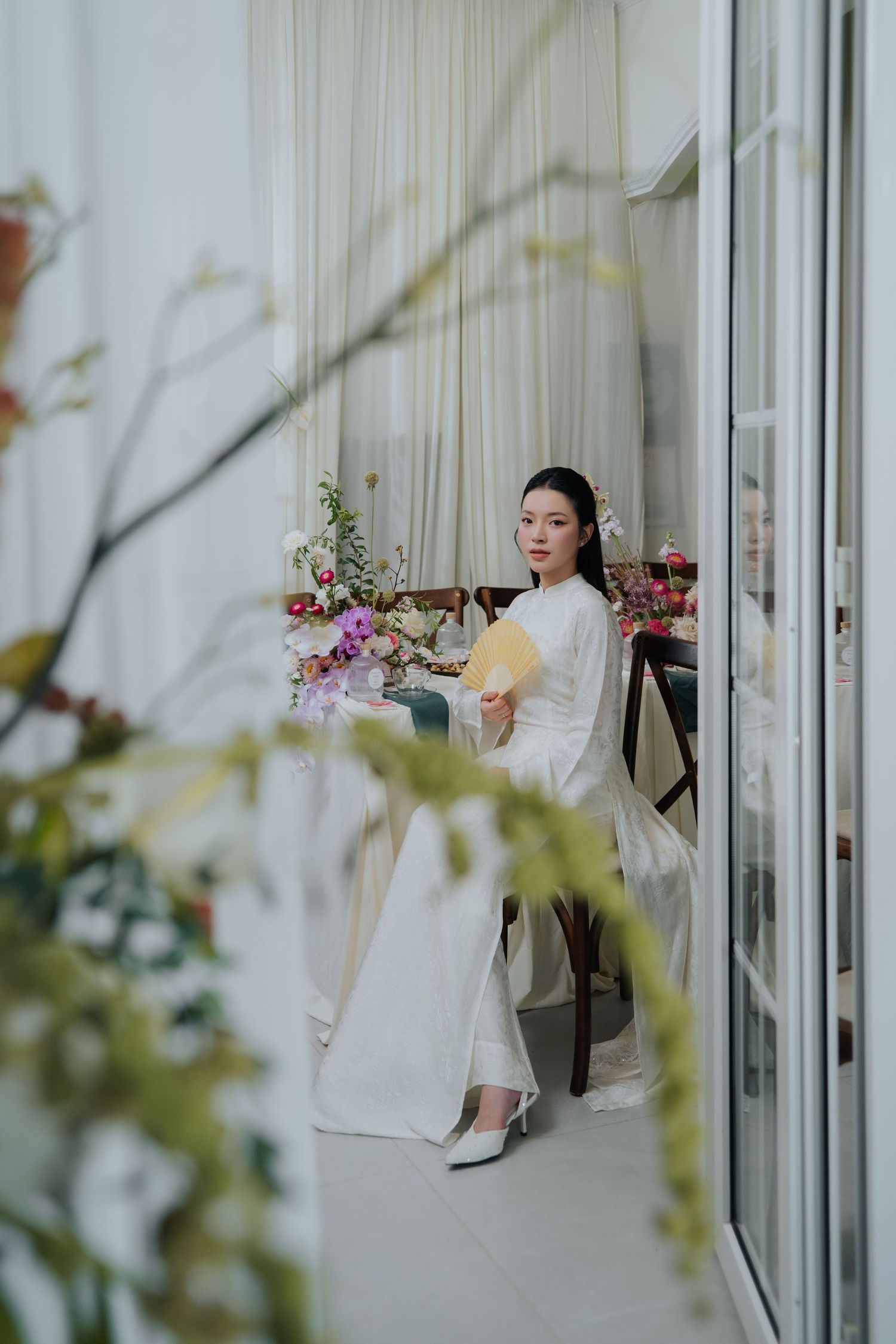 Elegant Vietnamese bride in white ao dai holding a fan seated by a floral arrangement near sheer curtained windows.