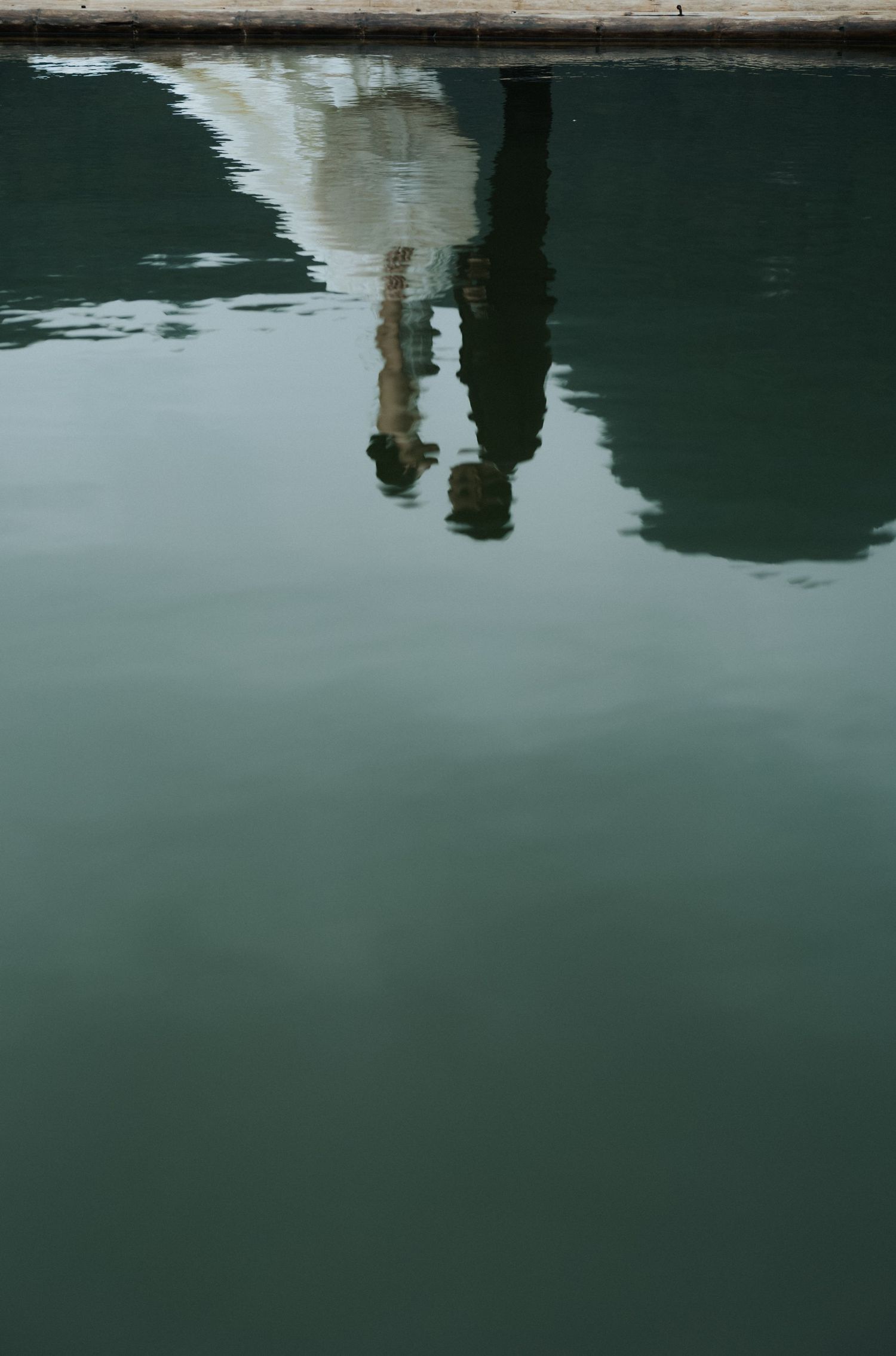 Reflection of a person standing on a dock mirrored in calm green water surface.