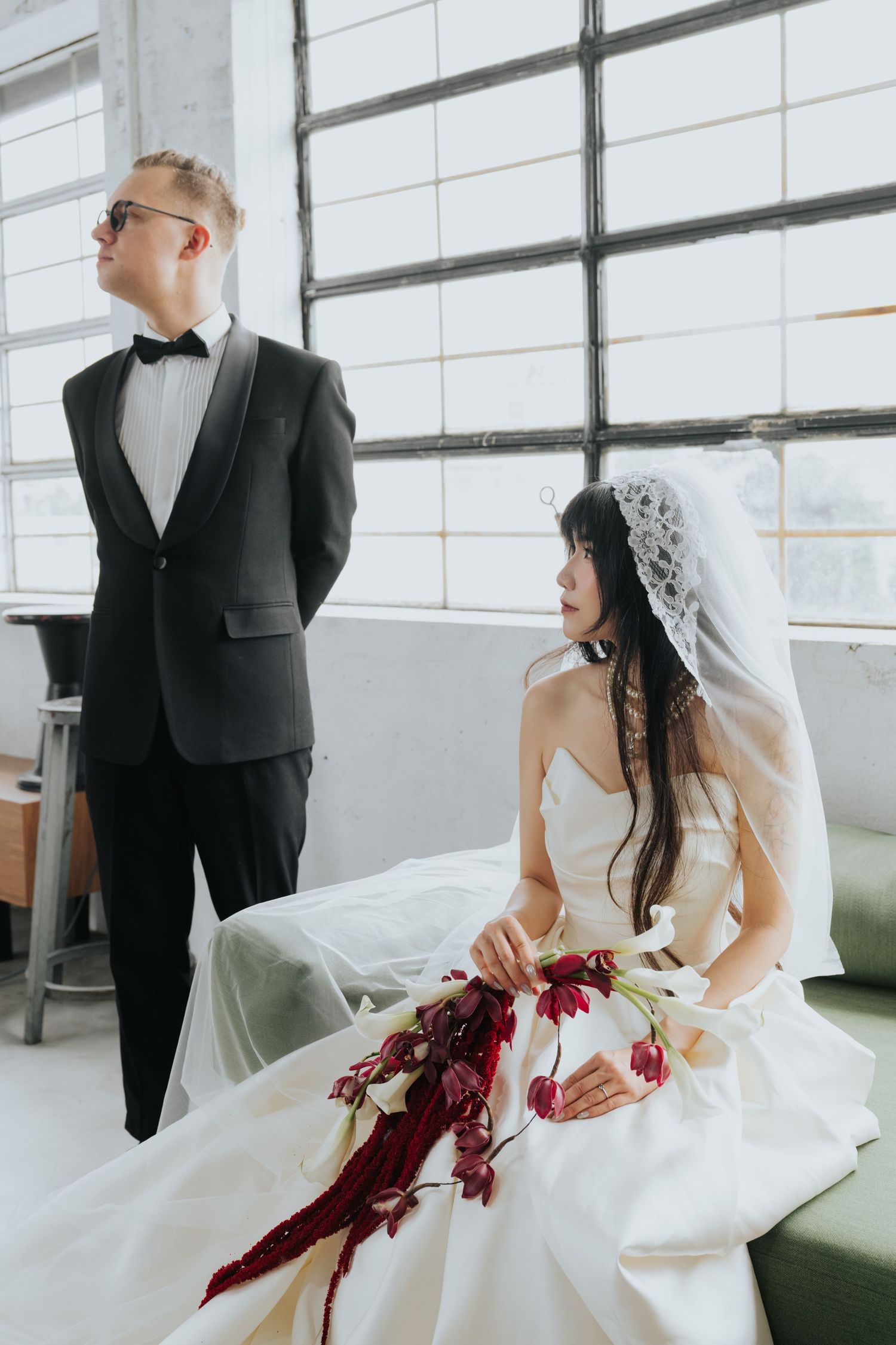 A bride in a white gown with veil holds red flowers while groom in black tuxedo stands nearby in an industrial venue.