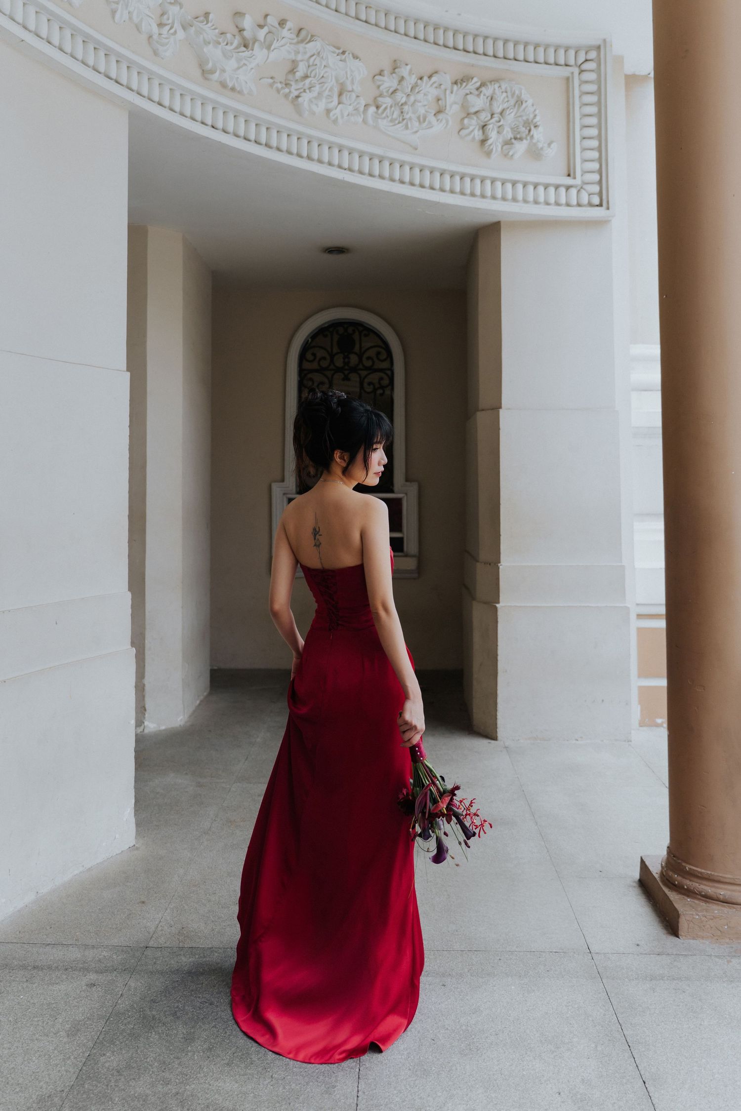 Woman in backless red satin gown holding bouquet, standing in grand white architectural corridor.