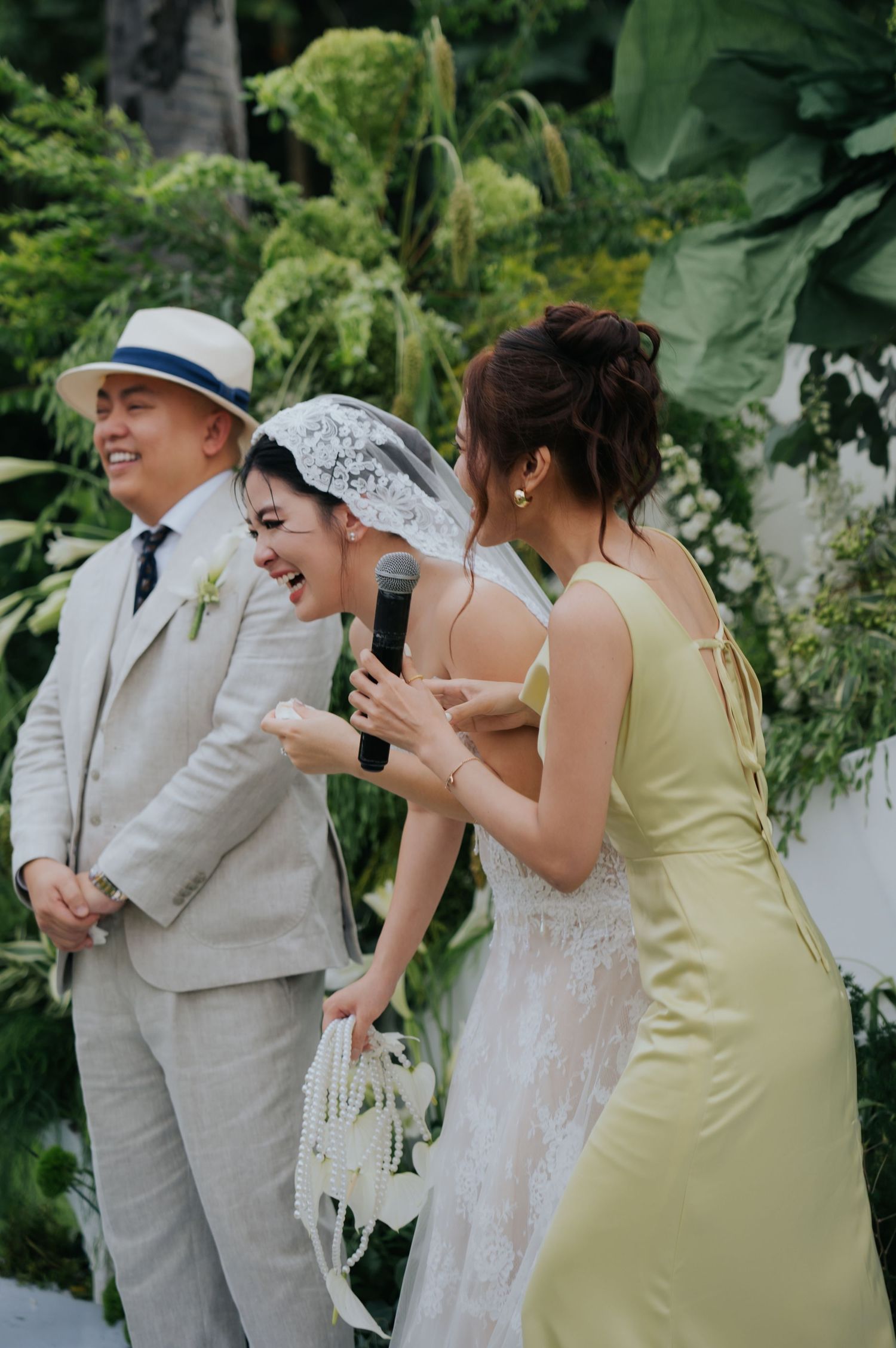 Bride in lace veil and groom in linen suit laugh with bridesmaid in yellow dress at outdoor tropical wedding ceremony.
