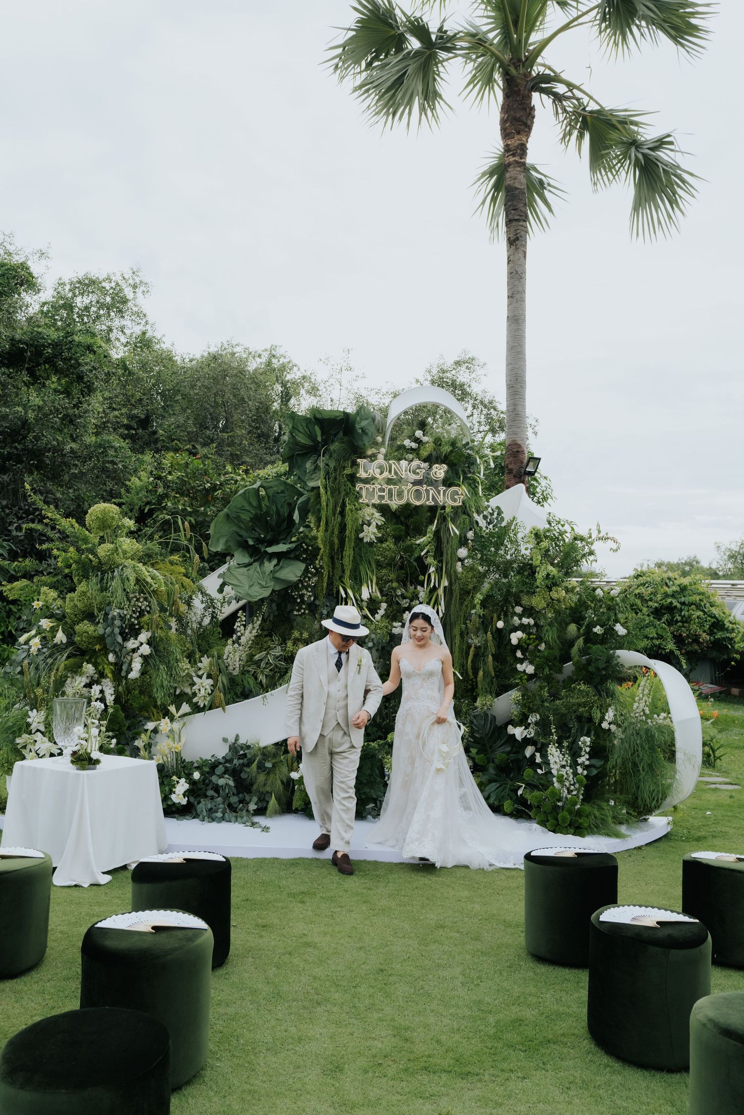 Bride and groom walk hand-in-hand at an elegant outdoor garden wedding ceremony with lush greenery backdrop.
