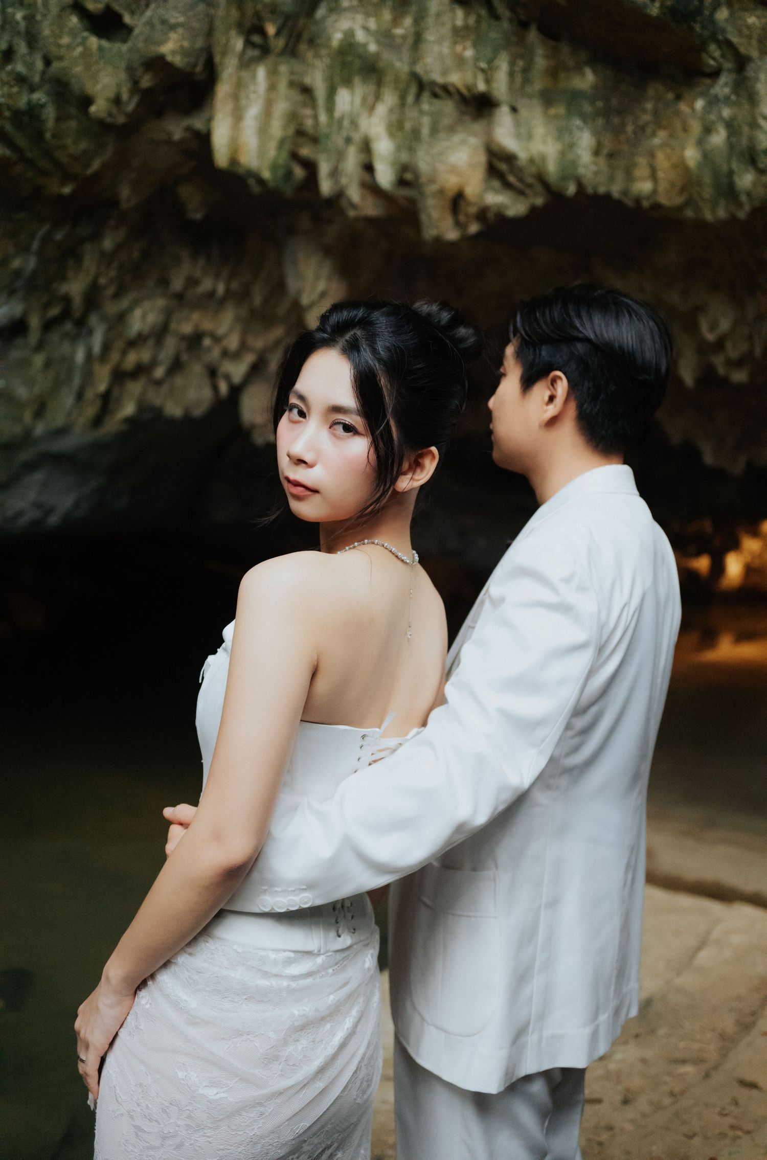 Elegant couple in white outfits standing in a cave, woman in backless gown glancing back softly.