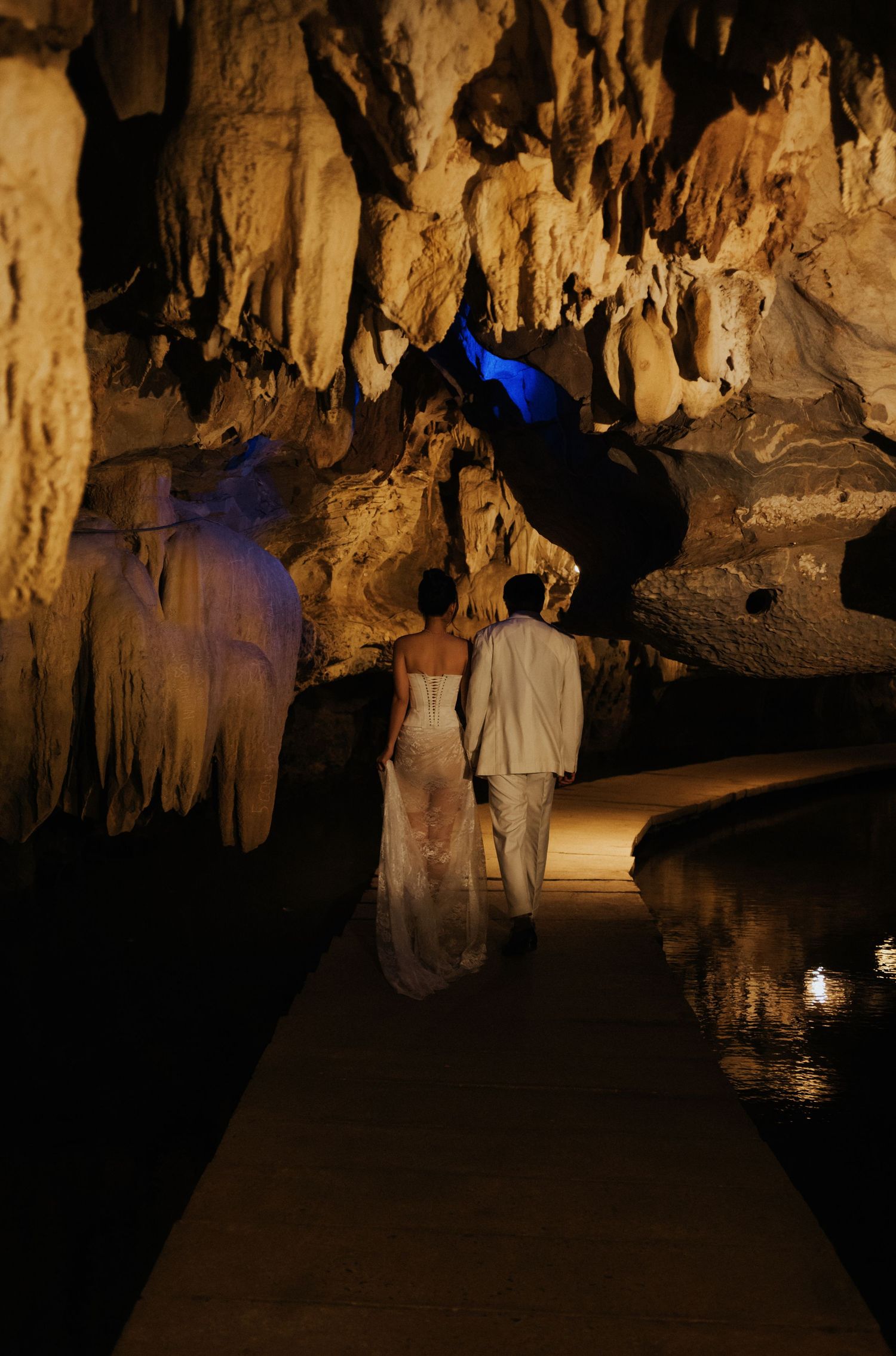Couple in wedding attire walking through an illuminated limestone cave with stalactites and a reflective pool.