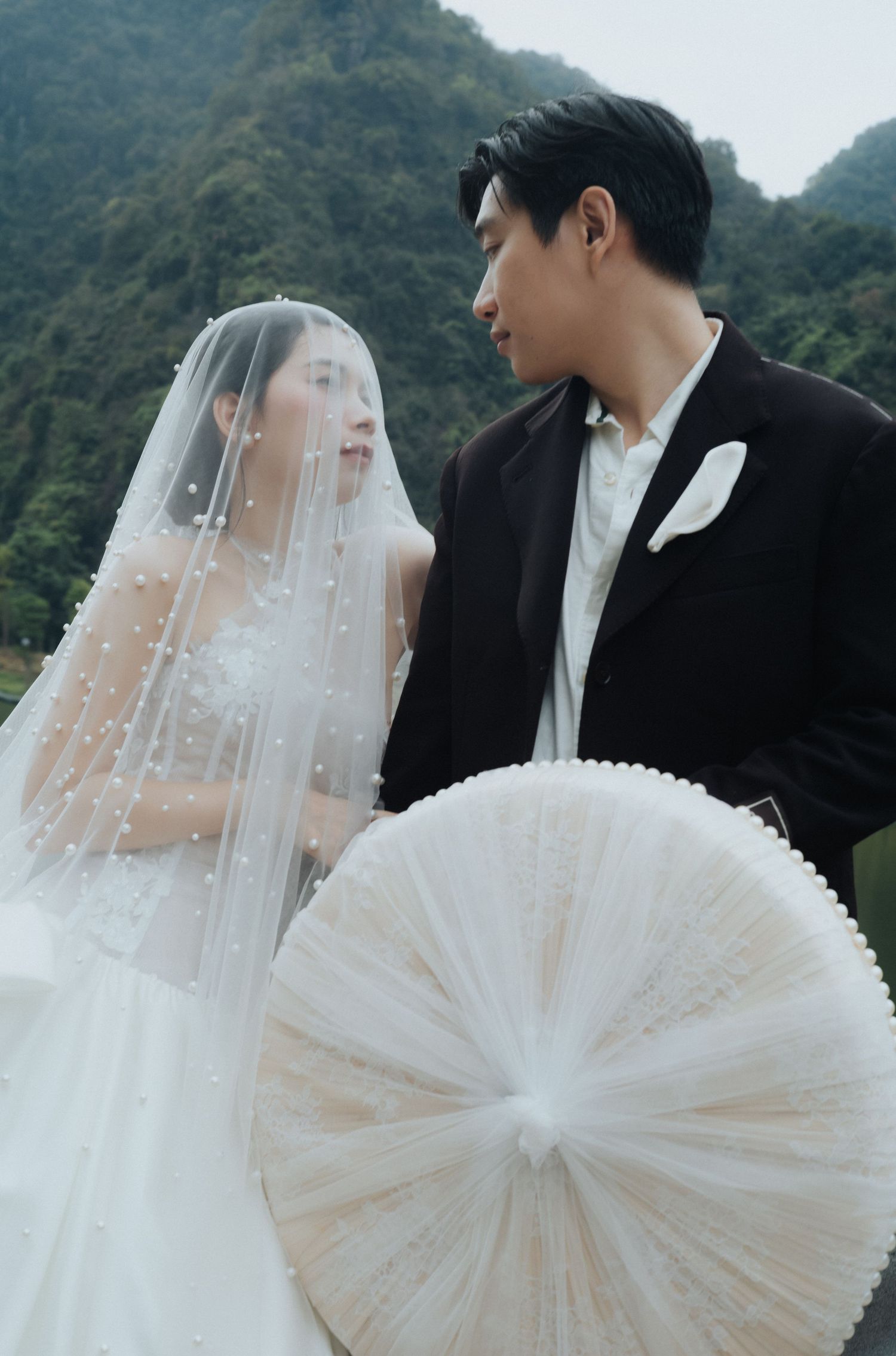 A bride in a pearl-adorned veil and groom in a black suit share a romantic moment with misty mountains behind them.