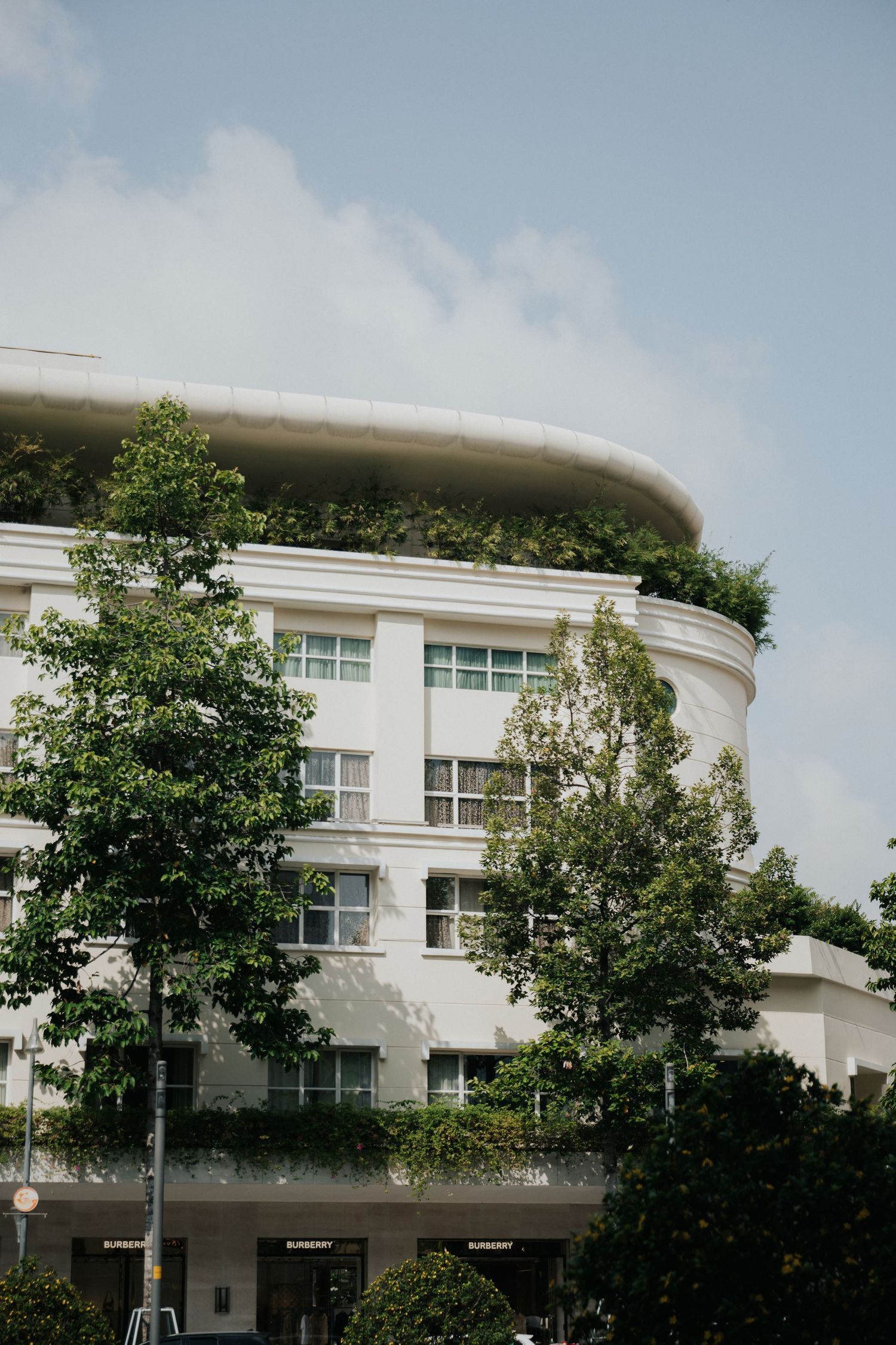 White curved building with lush green rooftop garden and tree-lined facade under a partly cloudy sky.