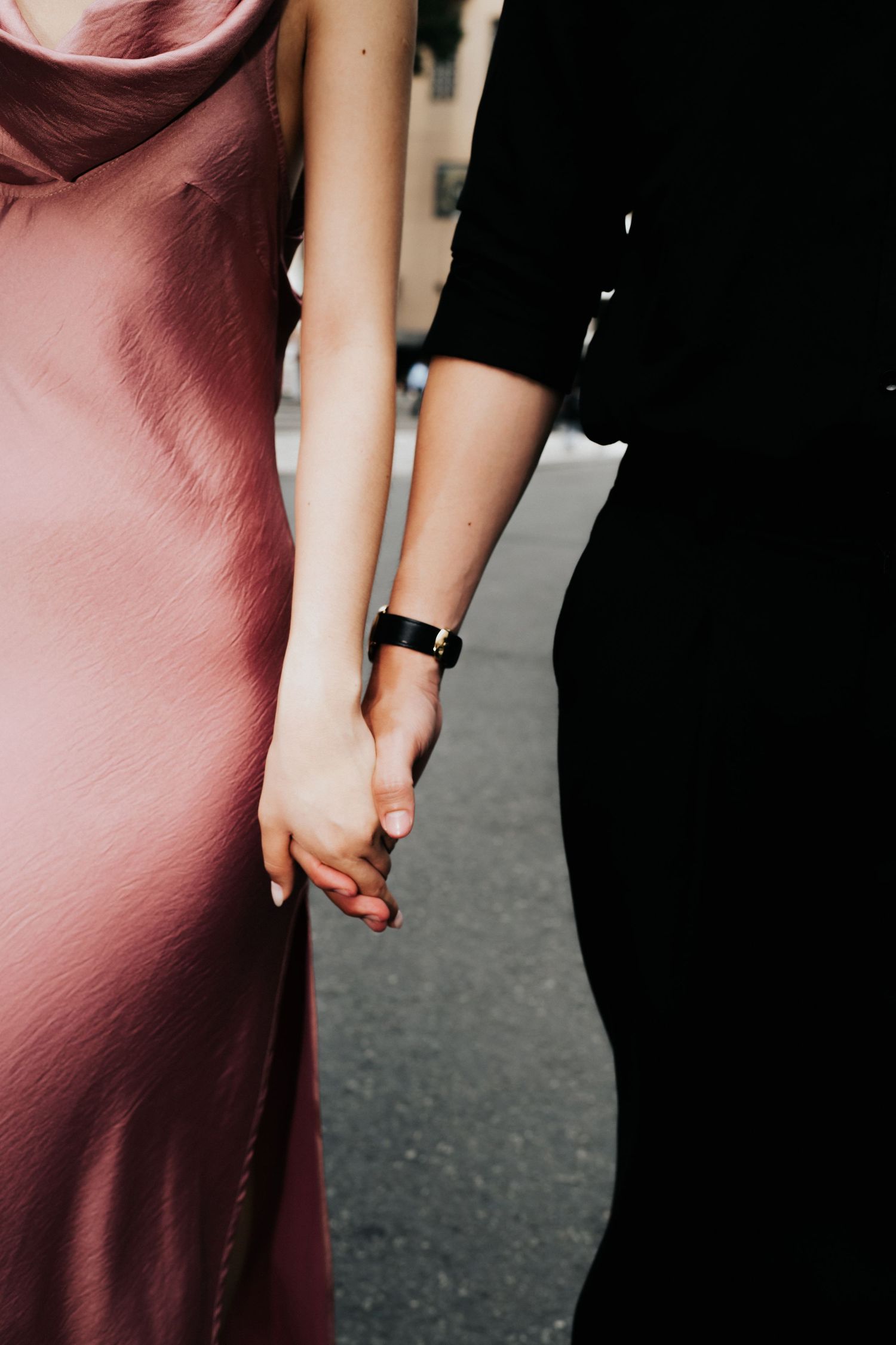 Two people holding hands, one wearing a pink satin slip dress and the other in black, with a black watch on their wrist.