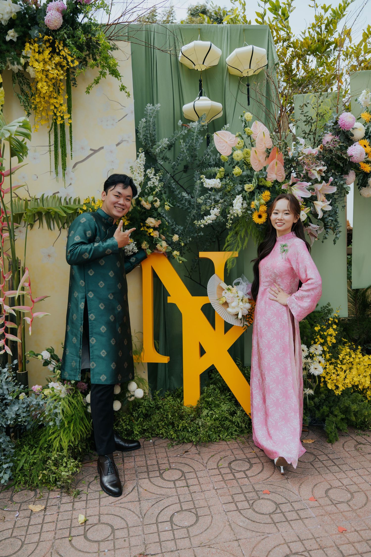 Couple in traditional Vietnamese ao dai attire posing before floral backdrop with decorative NK monogram letters.