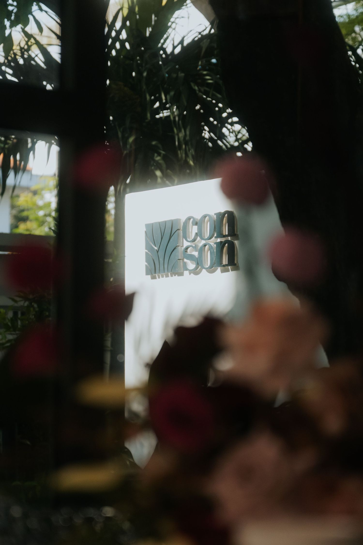 A glowing Con Son sign framed by tropical palms and blurred pink flowers in warm, moody light.