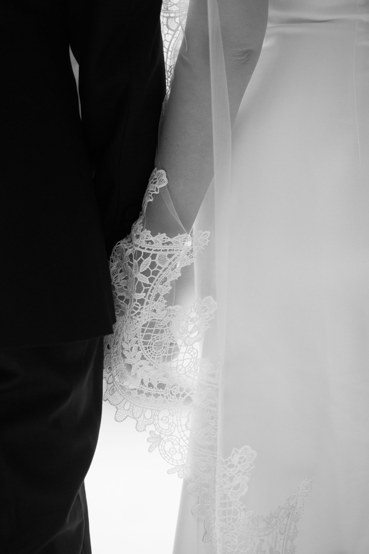 Black and white close-up of bride and groom holding hands, showcasing intricate lace wedding dress details and veil.
