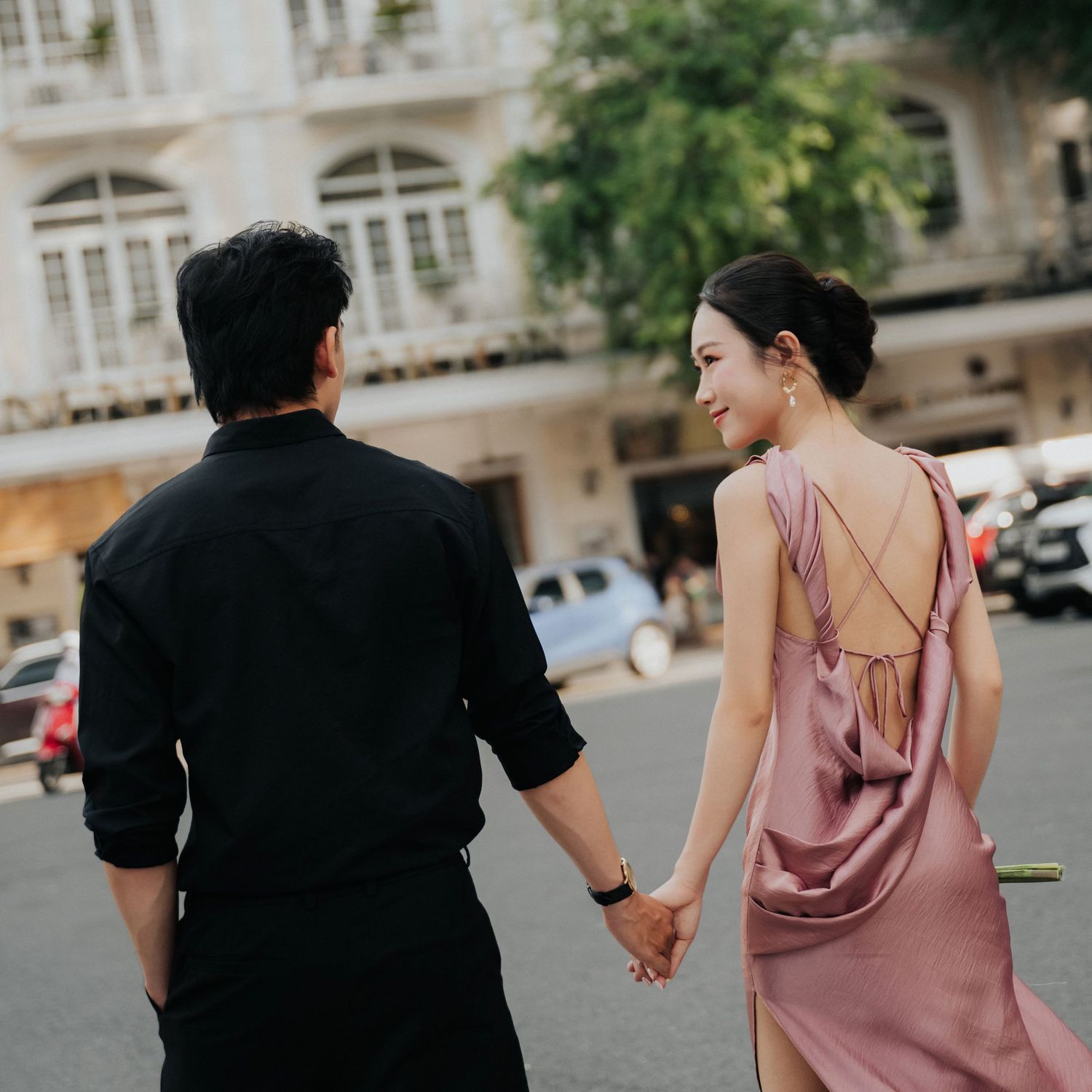 A couple holds hands while walking on a city street, the woman wearing an elegant backless pink satin dress.
