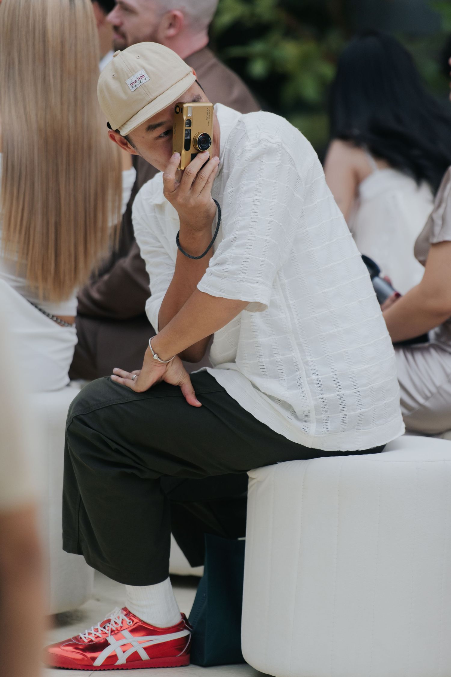 Person in white shirt, cap, and red Onitsuka Tiger sneakers sitting and taking a photo with a gold camera.