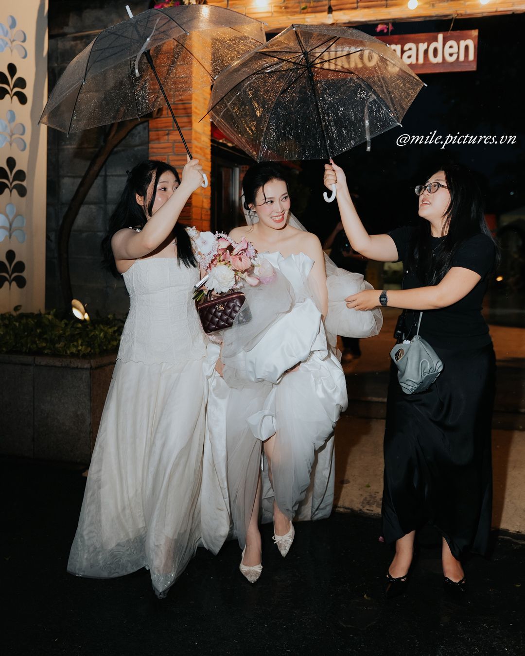 Two brides in white gowns hold umbrellas with a helper outside Nikko Garden restaurant on a rainy evening.