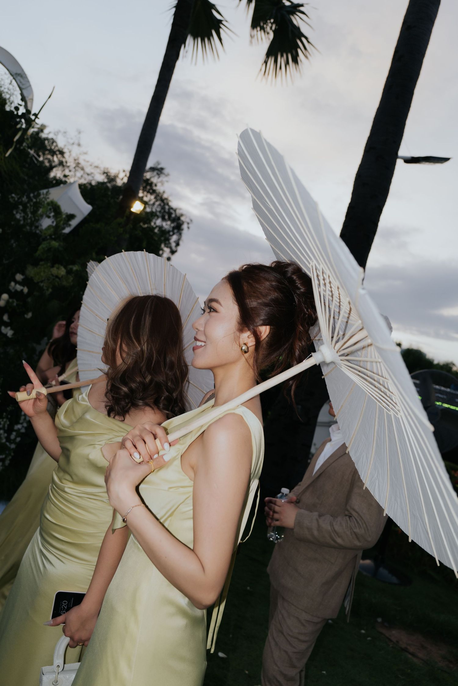 Two women in yellow dresses holding white parasols at an outdoor evening event with palm trees.