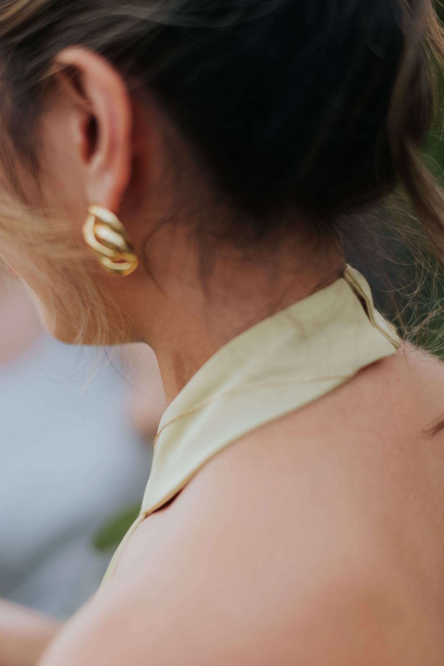 Woman wearing gold chain link earrings with light yellow halter top, hair up in bun, close-up side profile view.