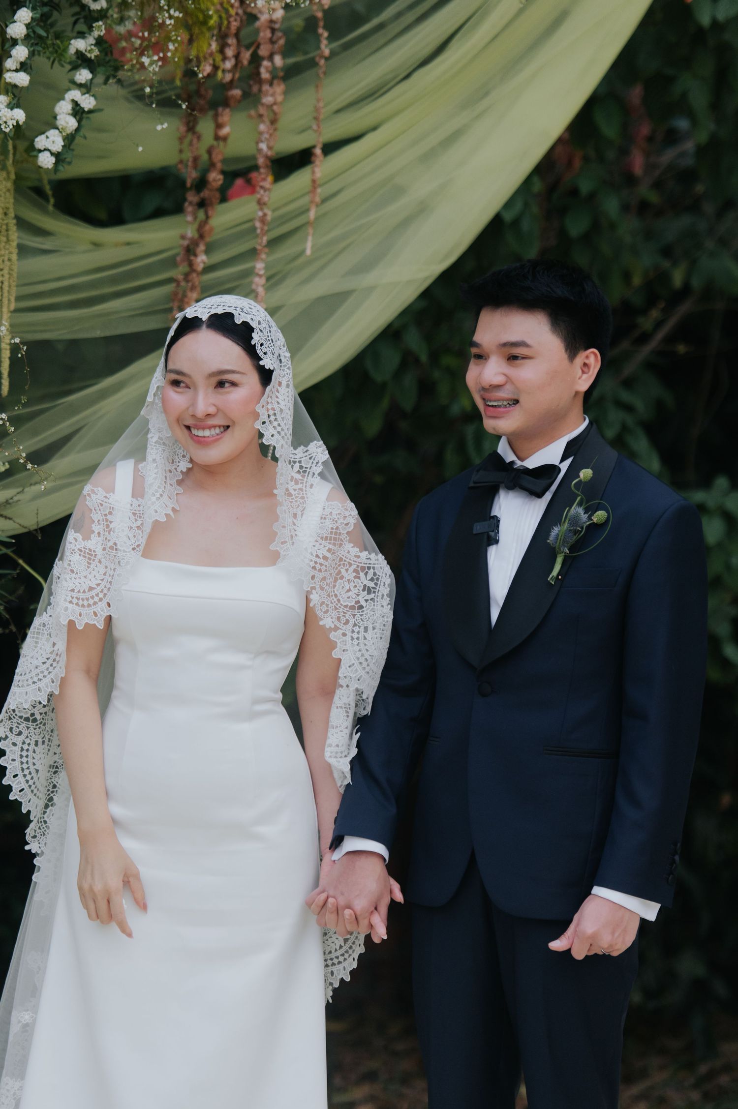 Smiling bride in white gown and lace veil holds hands with groom in navy tuxedo at outdoor wedding ceremony.
