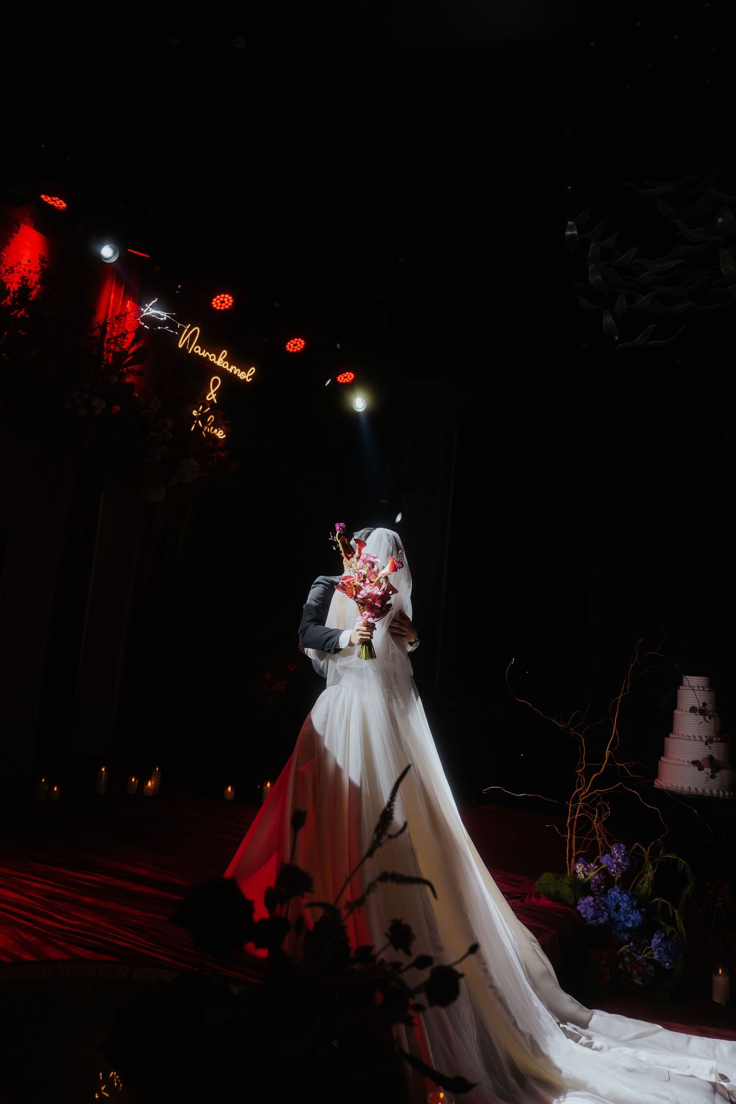 A bride in a white gown and veil holds a floral bouquet under dramatic spotlights at an elegant dark wedding venue.