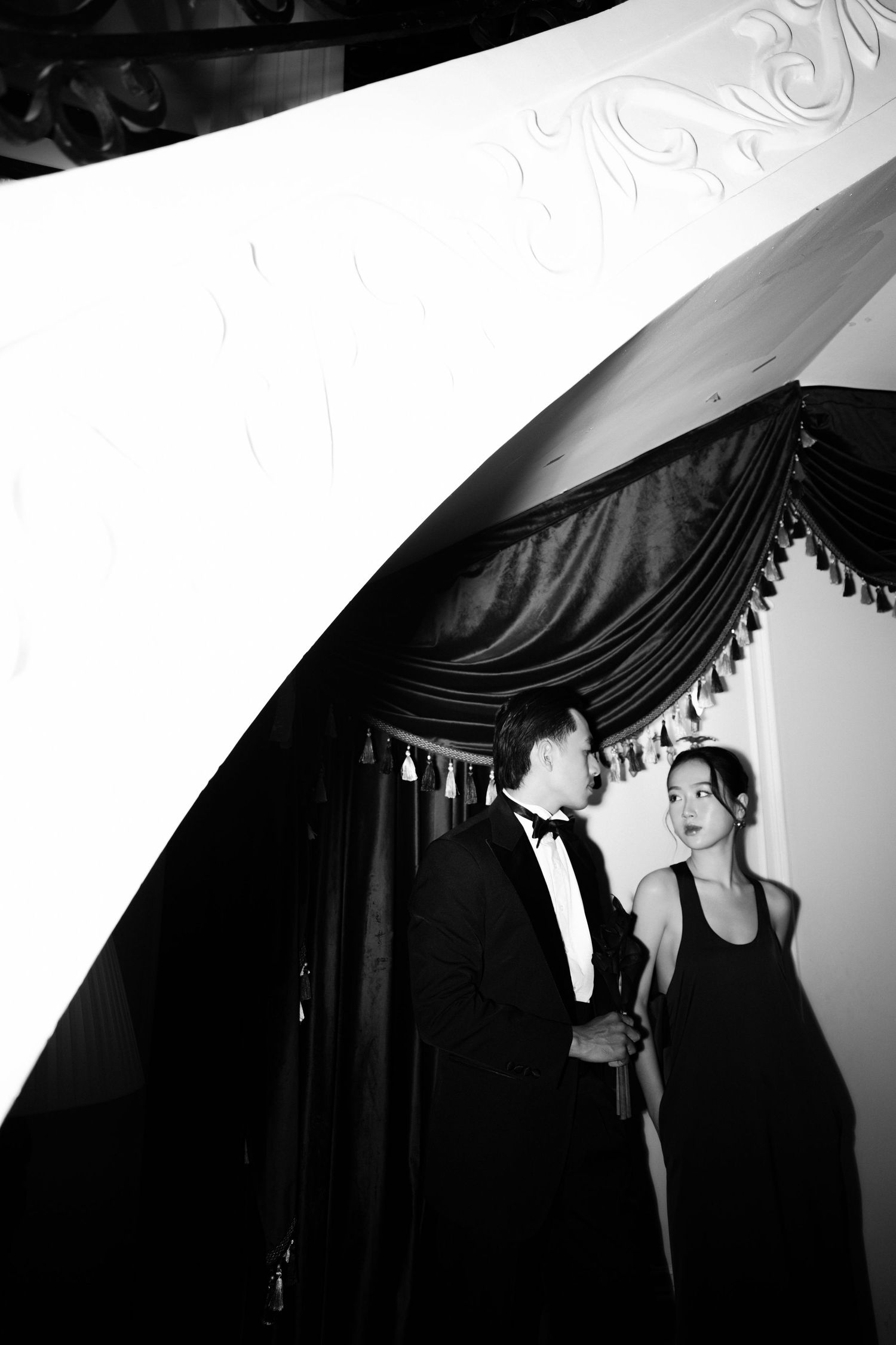 Black and white photo of elegant couple in formal attire conversing beneath ornate staircase with draped curtains.