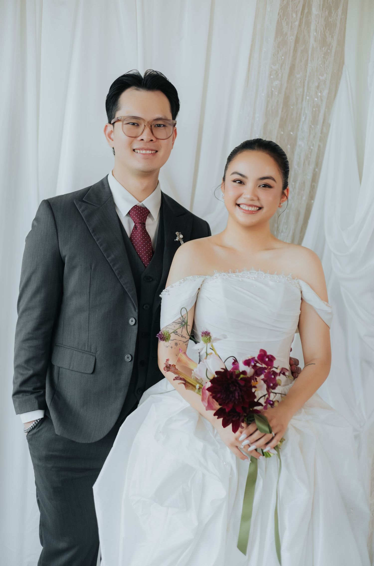 A smiling couple poses for a wedding portrait, groom in gray suit and bride in white off-shoulder gown holding a dark floral bouquet.