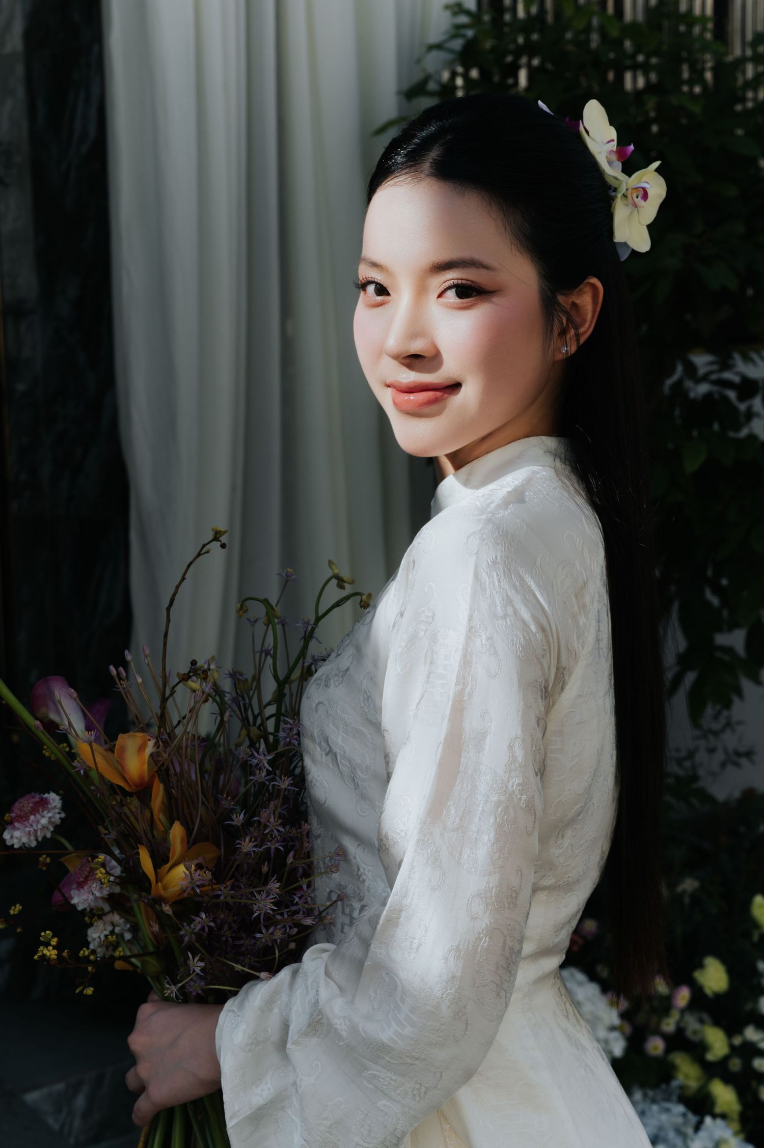 A young woman in a white floral dress holds a bouquet, smiling softly with a flower in her dark hair.
