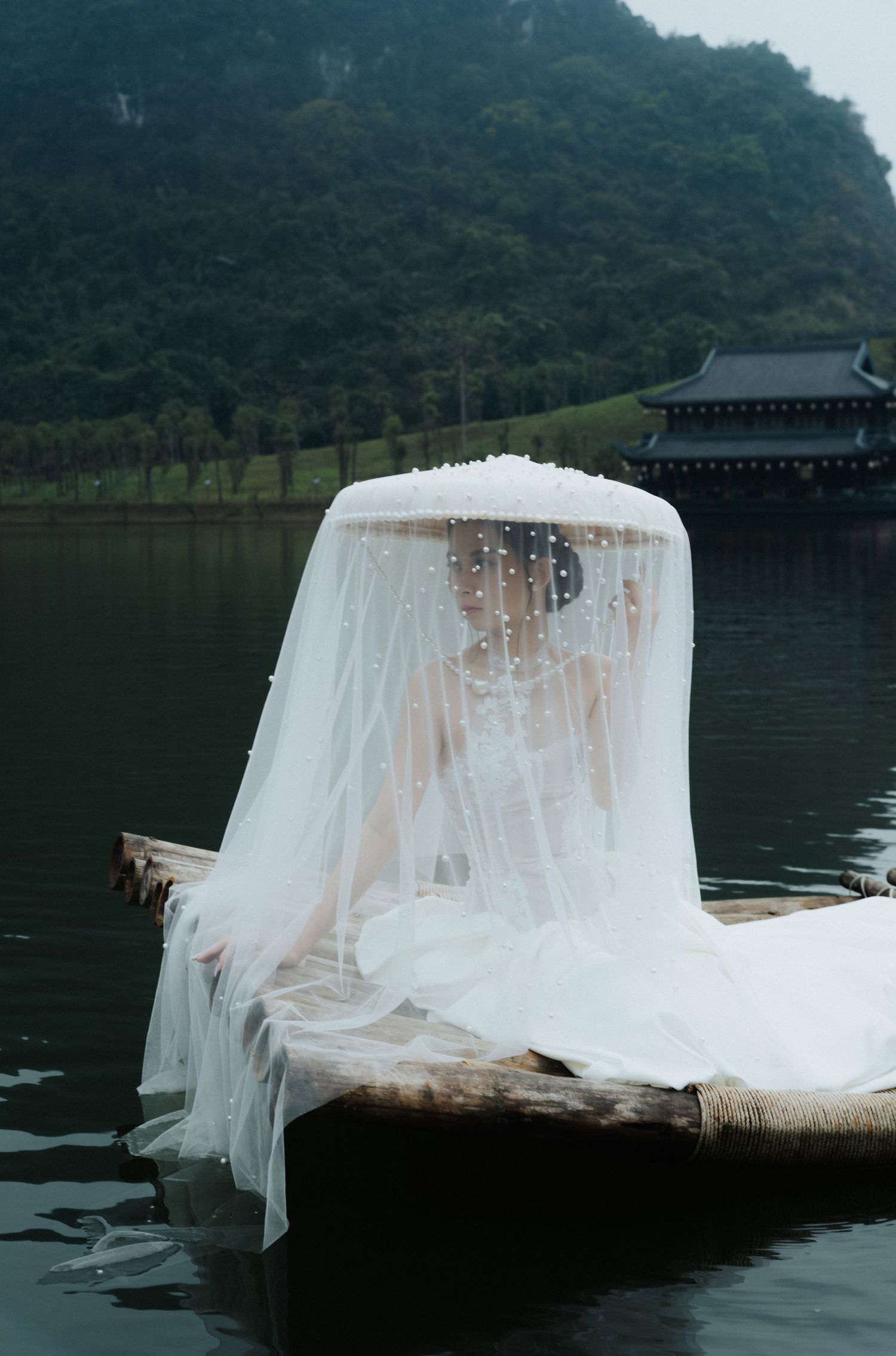 Bride in white wedding dress and veil sits on wooden boat on calm lake with lush green mountains in background.