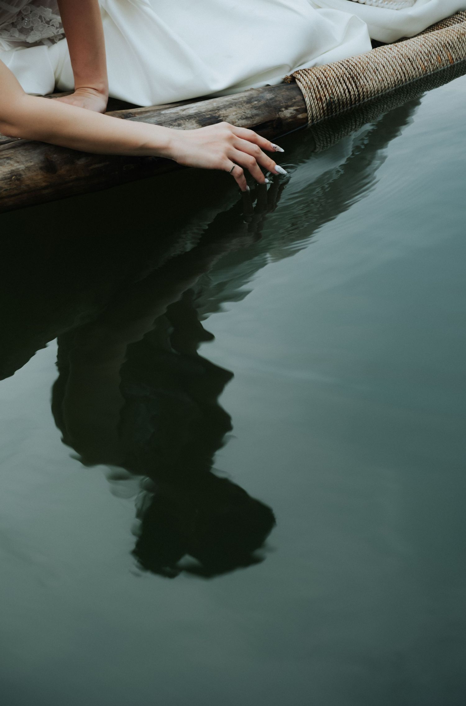 A woman in a white dress trails her hand in calm green water from a wooden boat edge.