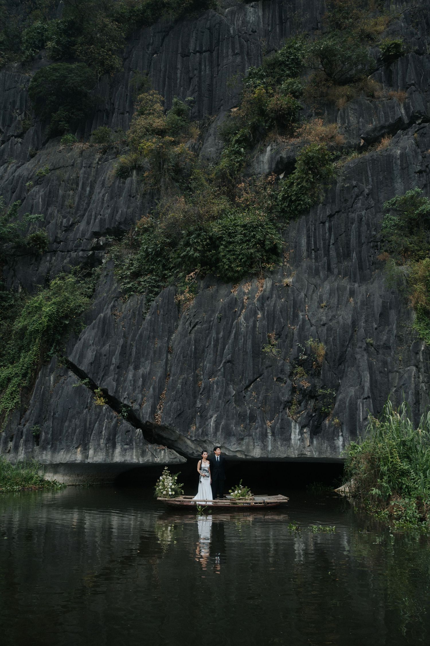 Couple standing on a boat before dramatic dark rock cliffs and lush greenery reflected in calm water.
