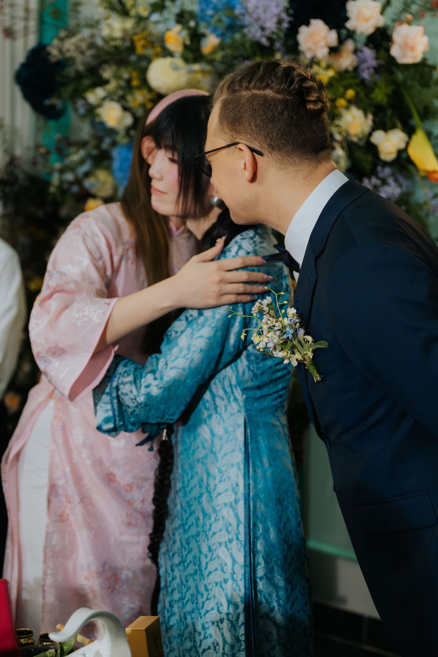 Groom in navy suit with boutonniere embracing woman in blue ao dai at floral wedding ceremony backdrop.