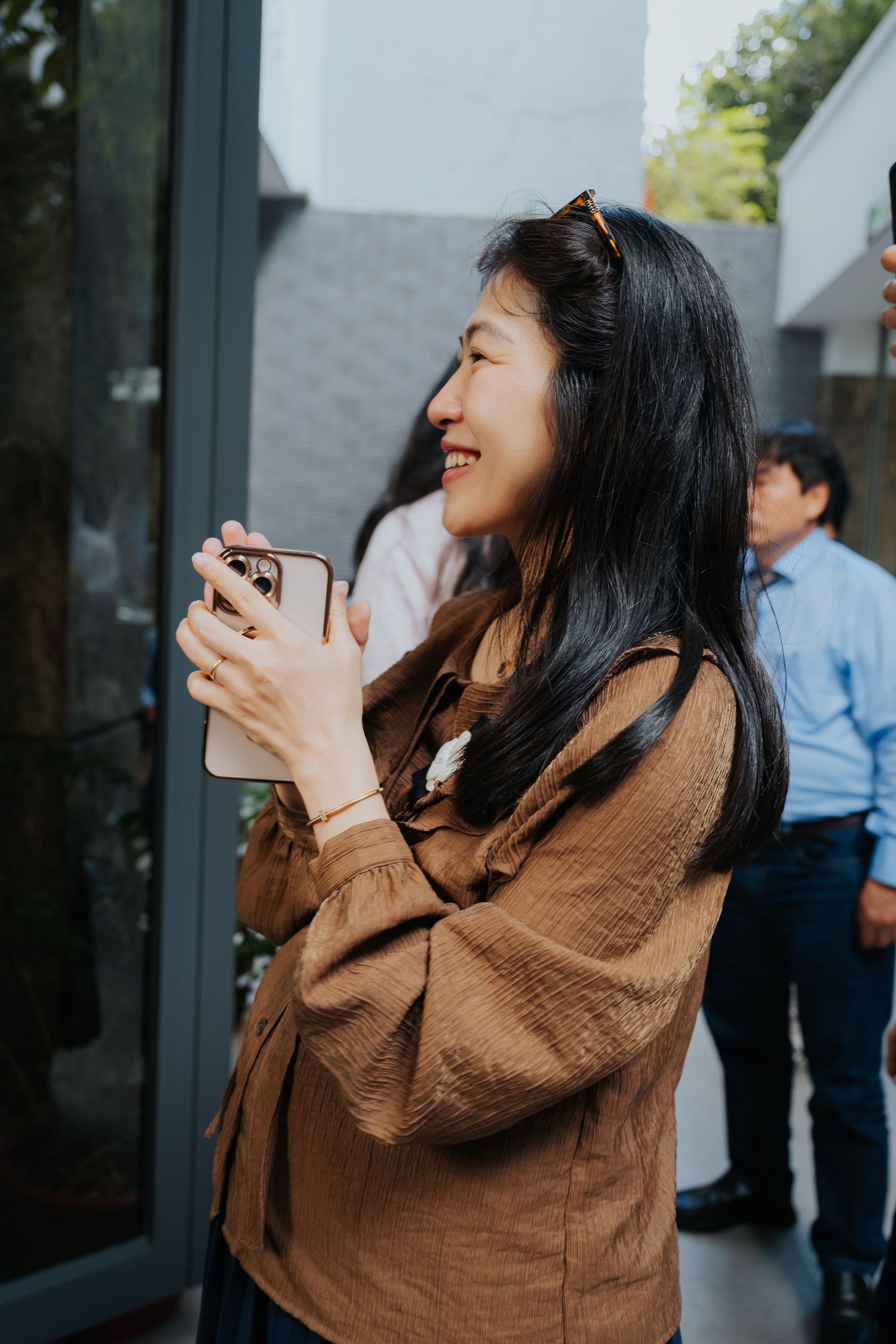 Smiling Asian woman in brown blouse holding smartphone outdoors at a social gathering.