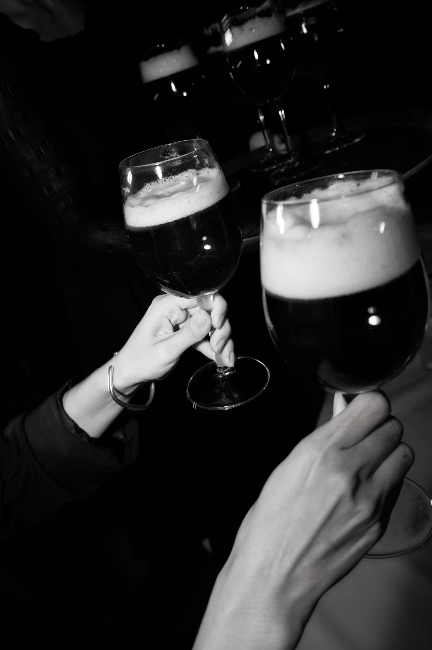 Two people toasting with dark beer glasses in a black and white photo, showing hands and frothy beverages.