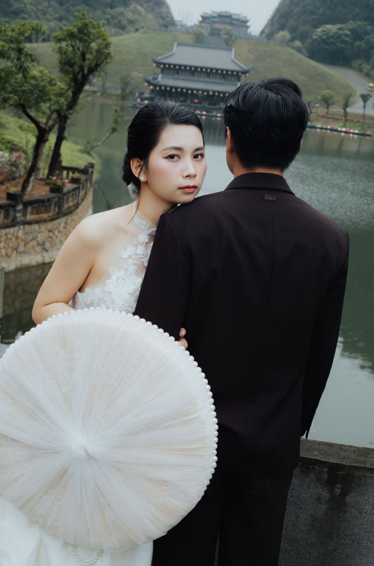 Asian bride in white gown holds parasol beside groom in black suit near scenic pagoda lake.