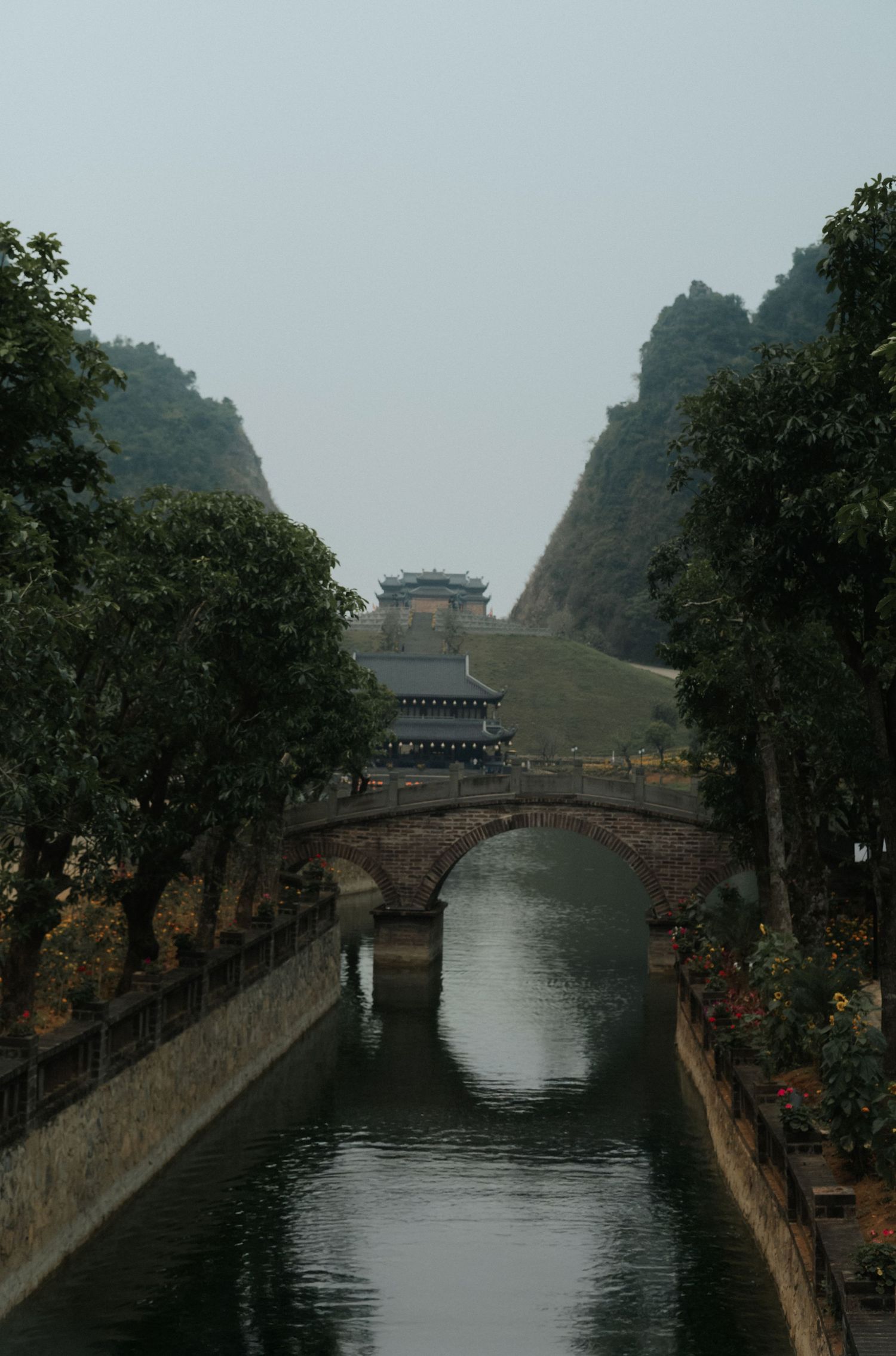 Ancient Chinese stone arch bridge over a serene canal leads to a traditional pagoda amid misty karst mountains.