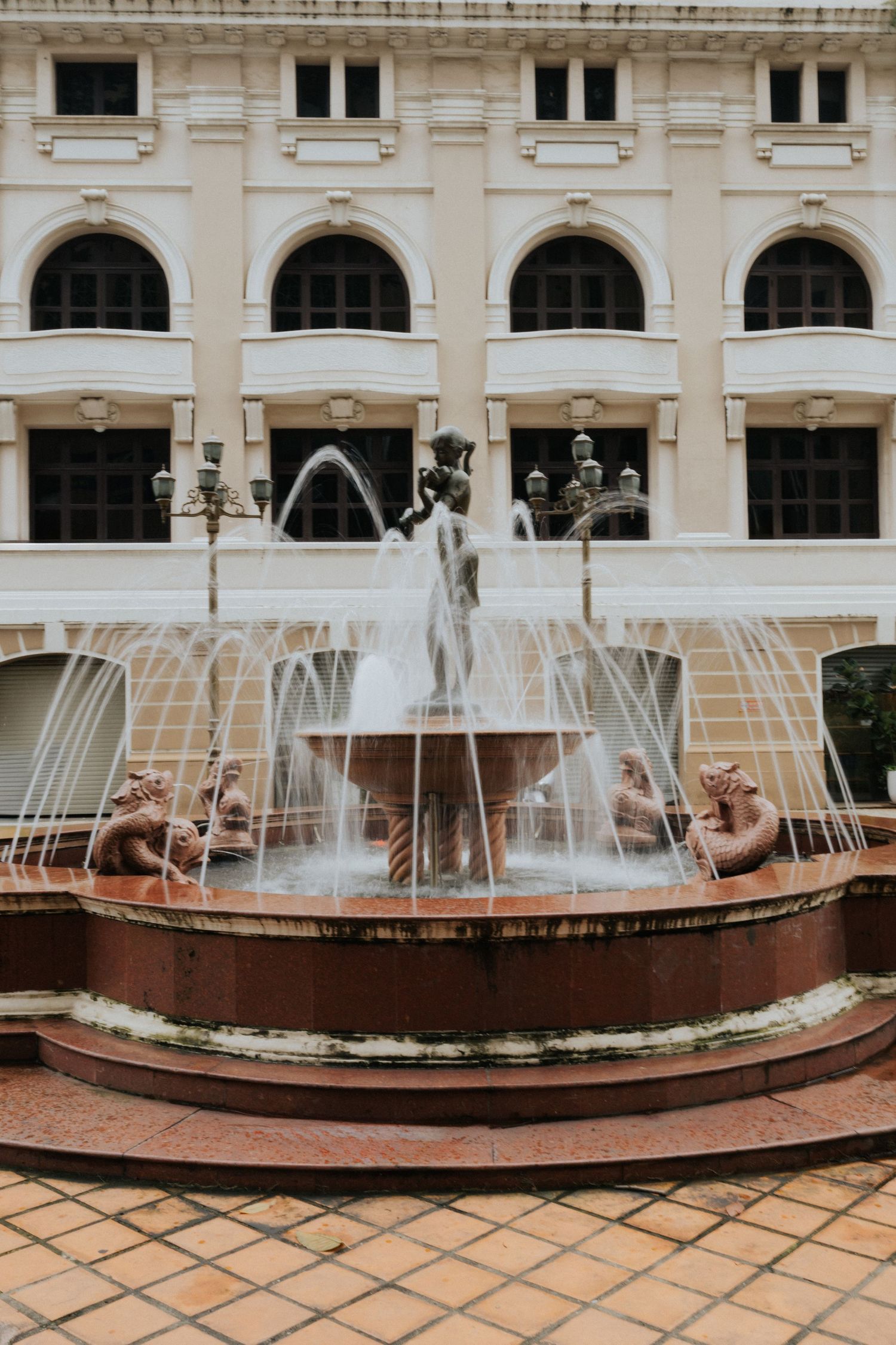 Ornate fountain with bronze sculptures in front of a classical colonial-style building with arched windows.