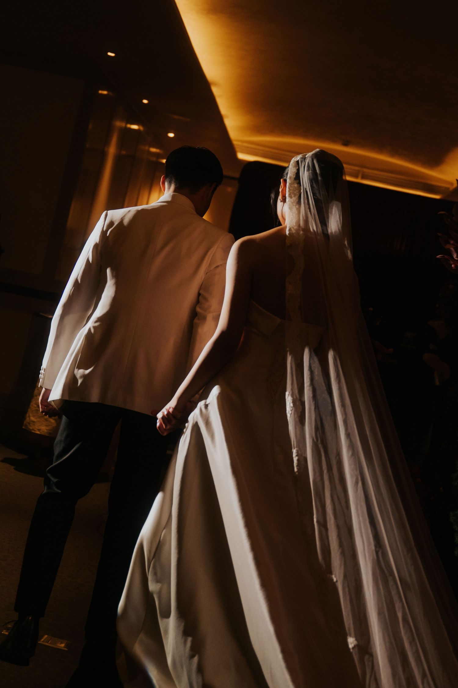 Bride in white gown and veil walks hand-in-hand with groom in white jacket, viewed from behind in dimly lit venue.