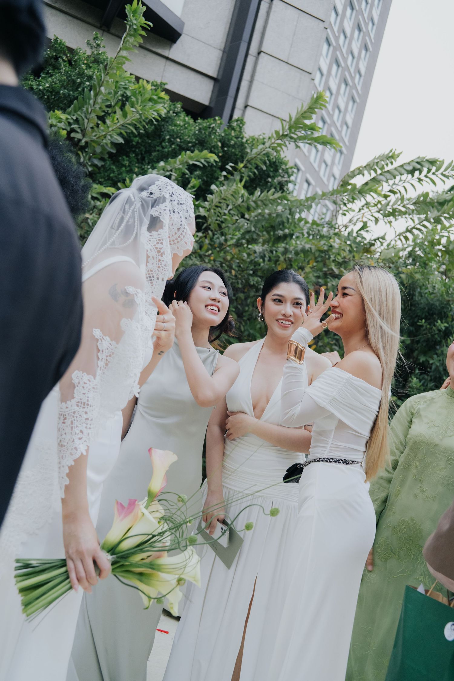 Bride in lace veil holding calla lily bouquet laughing with bridesmaids in white dresses outdoors.