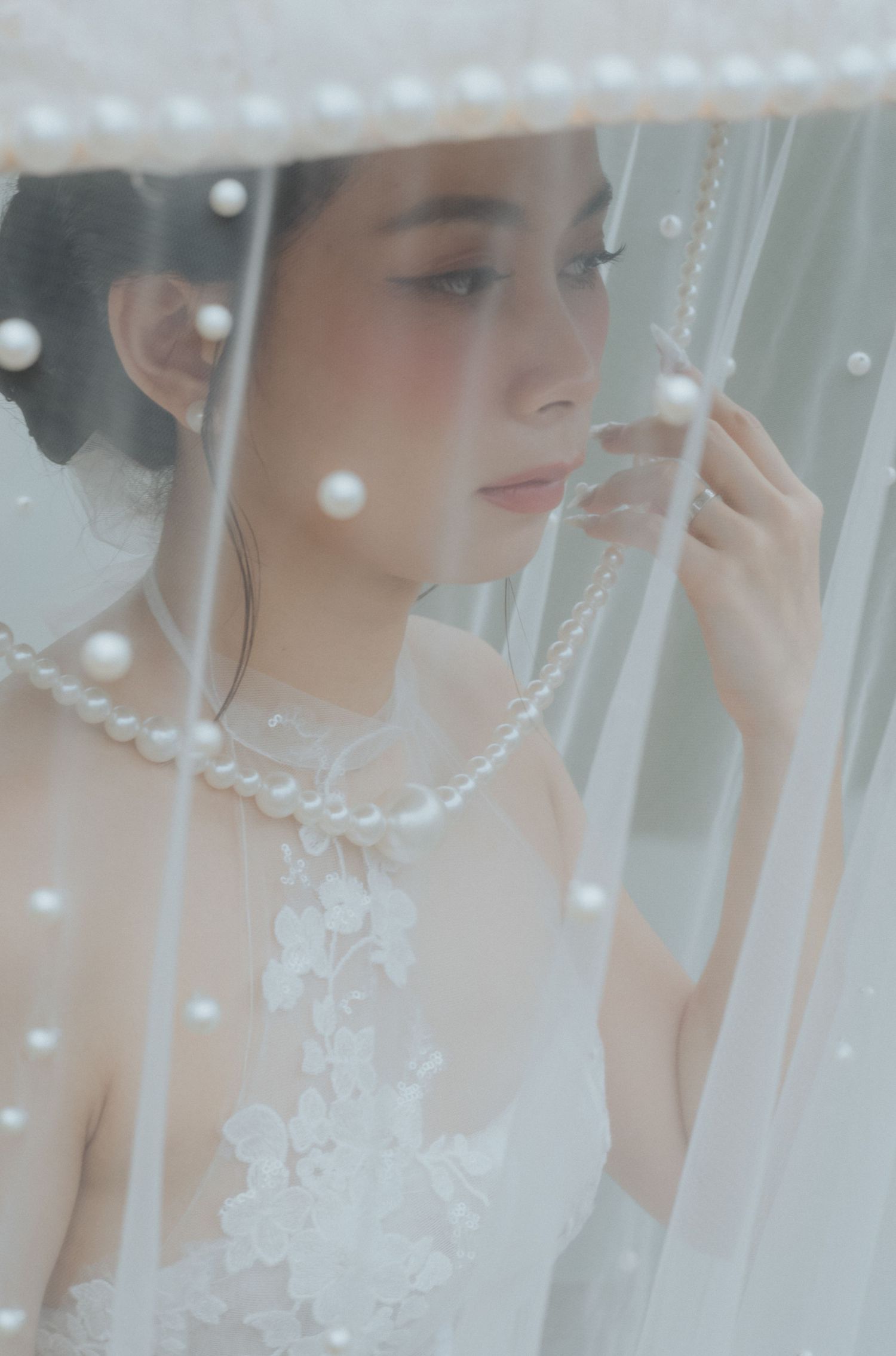 A bride in a white lace gown and pearl necklace gazes softly through a sheer veil adorned with pearl accents.