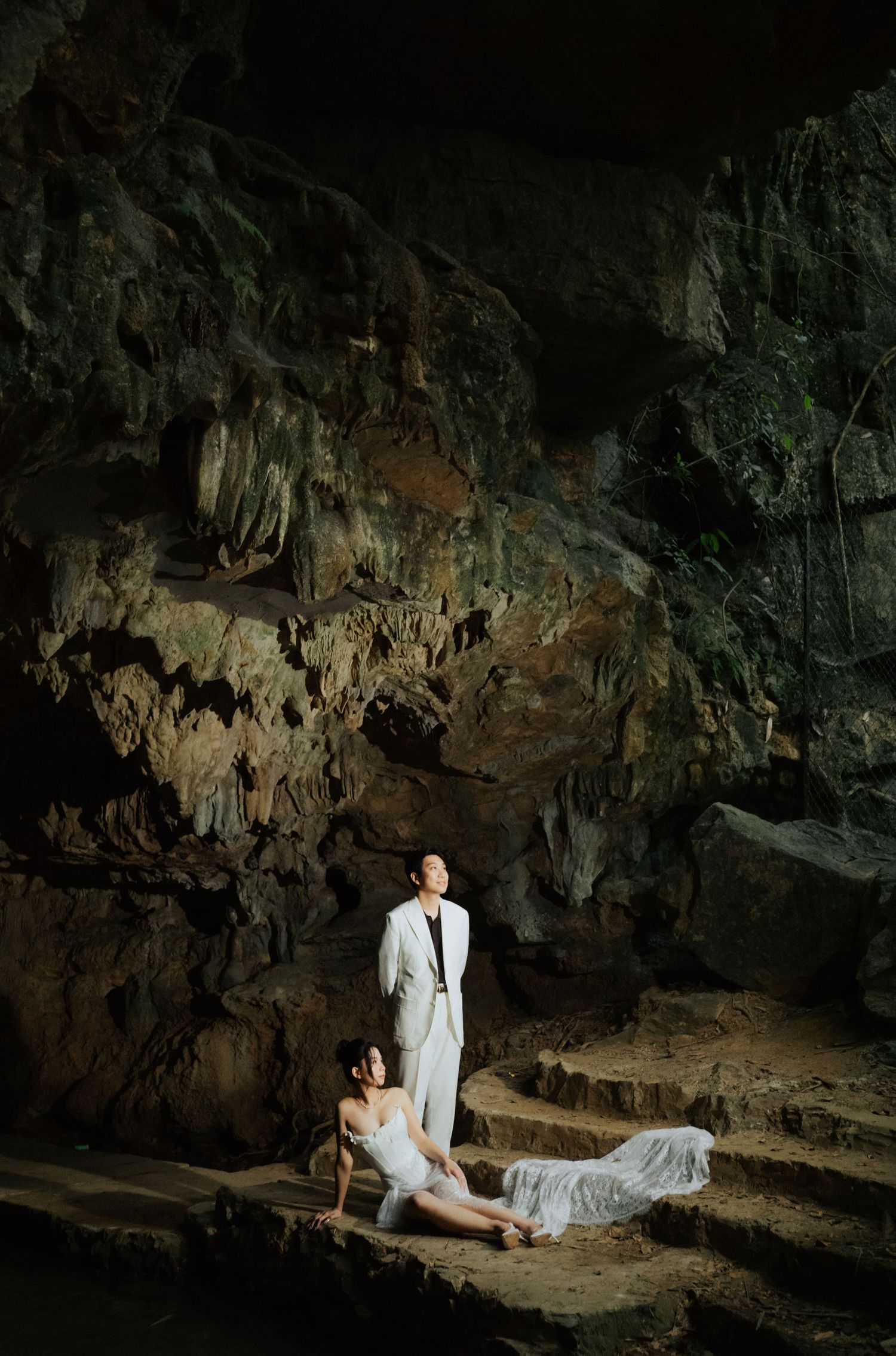 A couple in white wedding attire poses dramatically inside a cave with stunning rock formations and natural lighting.
