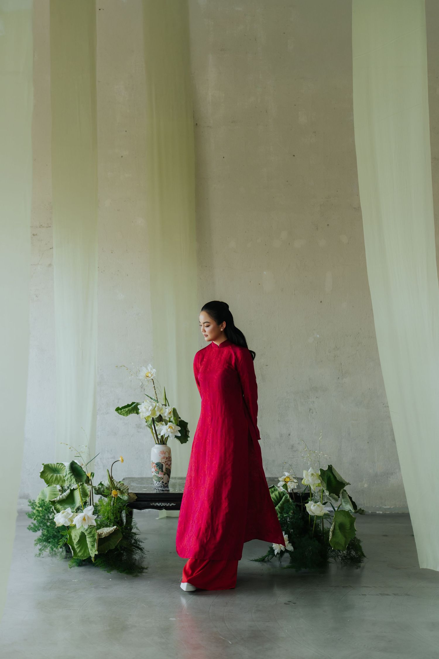 Woman in red áo dài gown standing near floral arrangements with white flowers and green leaves in a minimalist space.