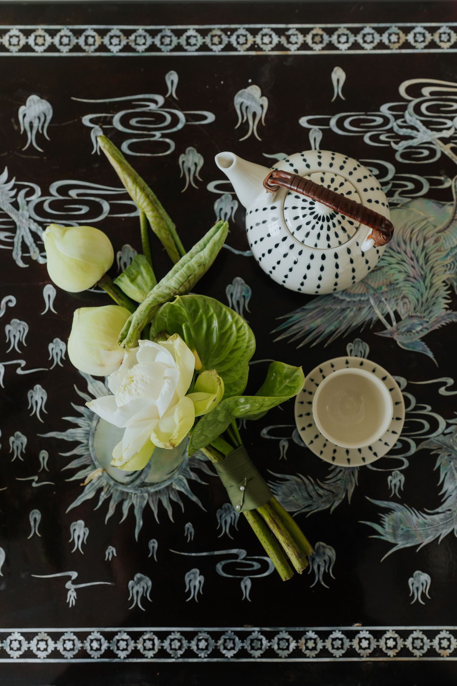 Black lacquer tray with white teapot, tea cup, and white lotus flowers with green leaves in an elegant Asian-inspired arrangement.