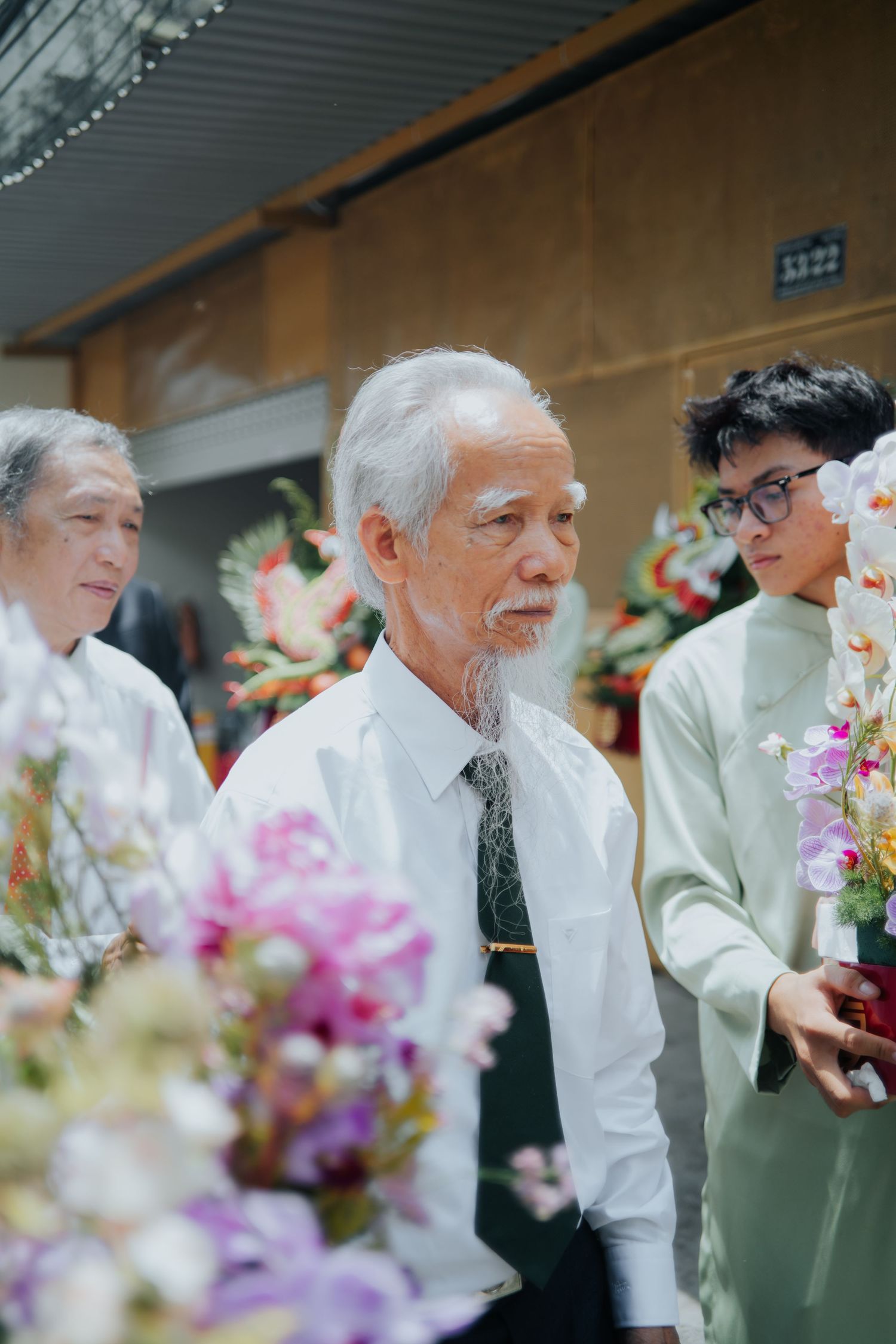 Elderly Vietnamese man with white beard in white shirt at a floral ceremony with family members.