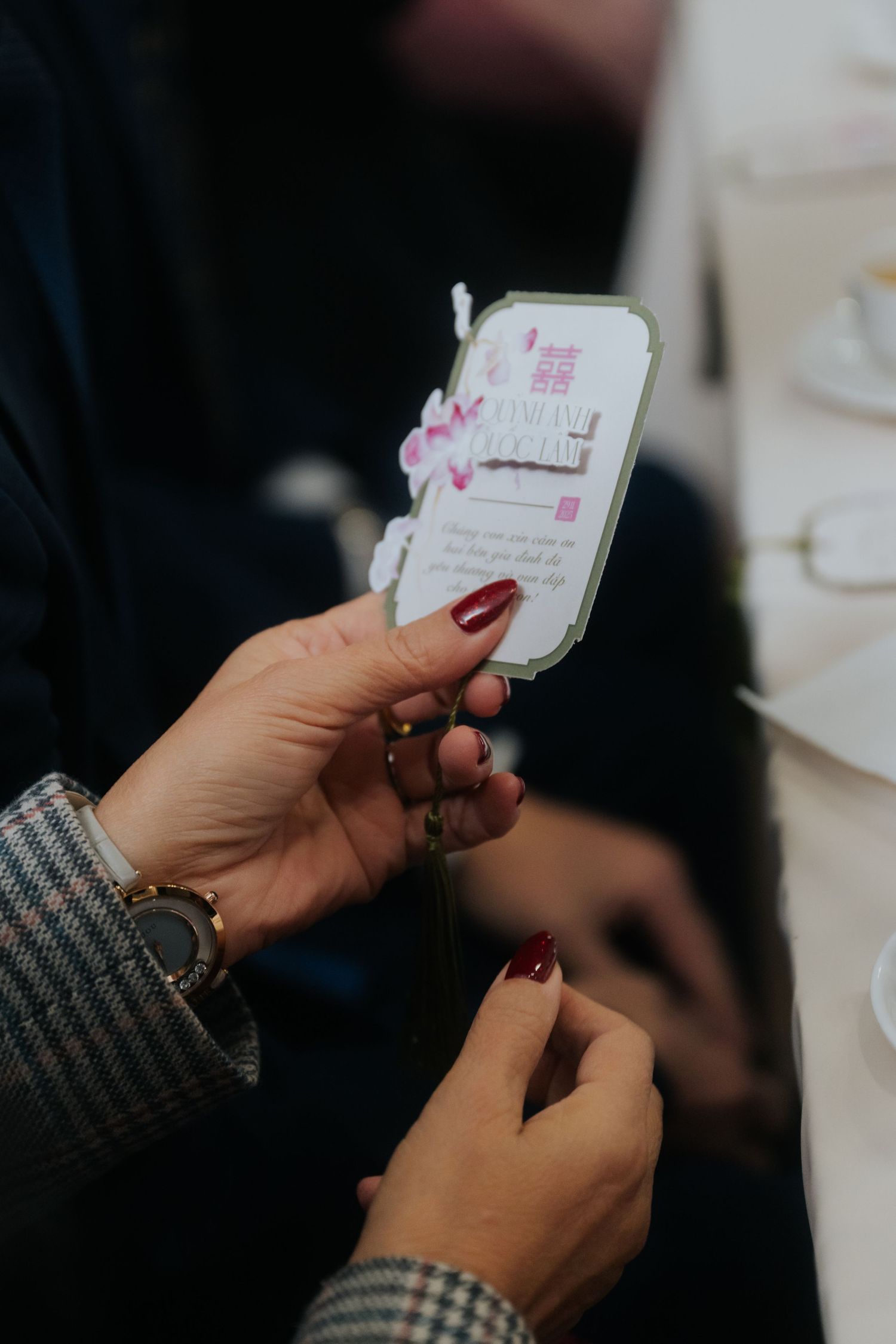 Hands with red nails holding a decorative Chinese wedding card with pink floral details and double happiness symbol.