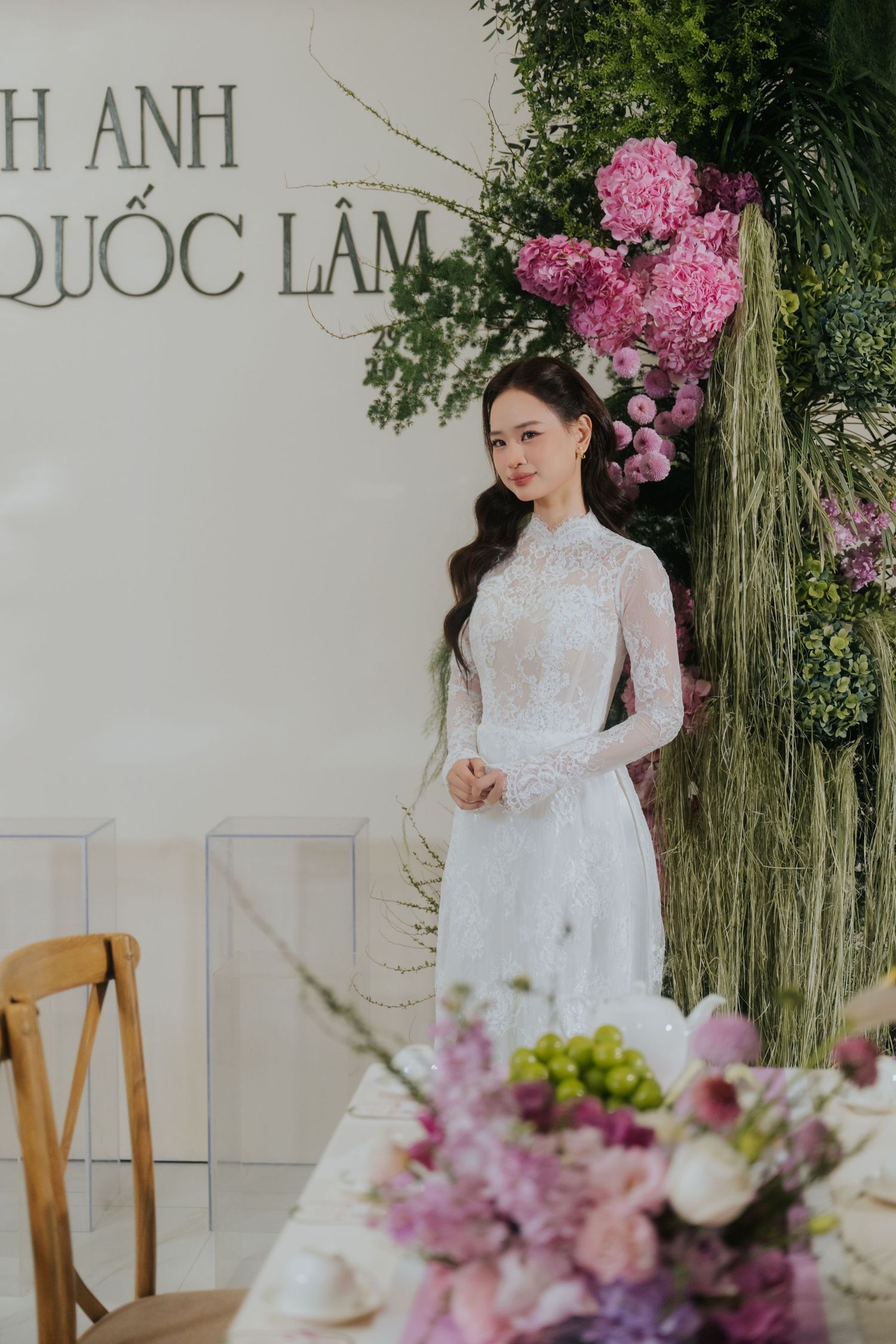 A bride in a white lace ao dai gown stands before a floral backdrop at a Vietnamese wedding ceremony.