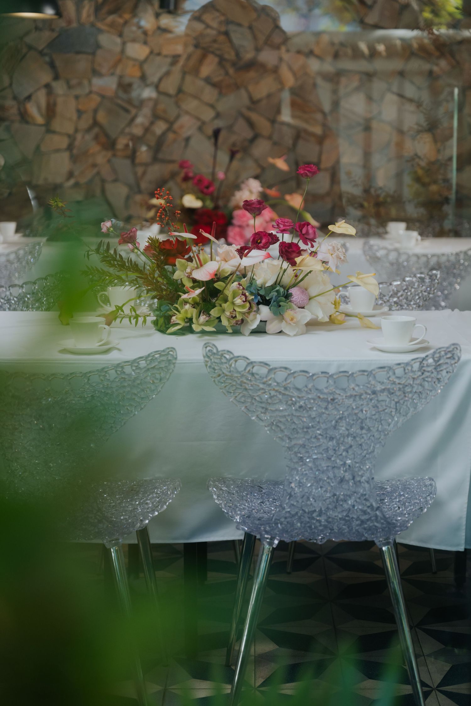 Elegant wedding table setting with colorful floral centerpiece, white linen, and transparent acrylic chairs against a stone wall.
