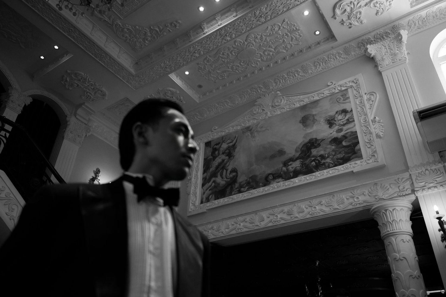 Black and white photo of a young man in a tuxedo with bow tie standing in an ornate ballroom.