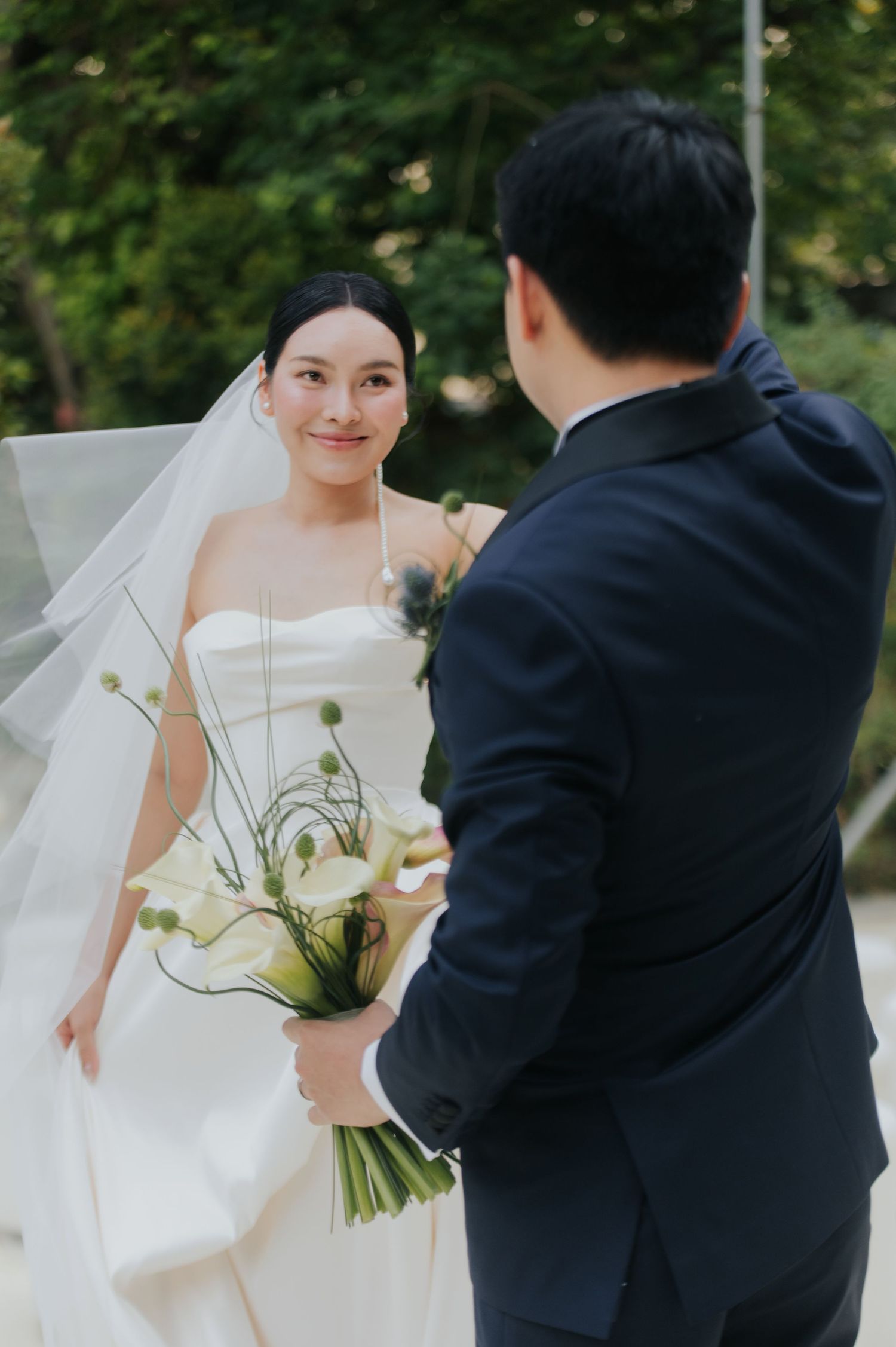 A bride in a white strapless gown and veil holds a bouquet of calla lilies, smiling at a groom in a navy suit.