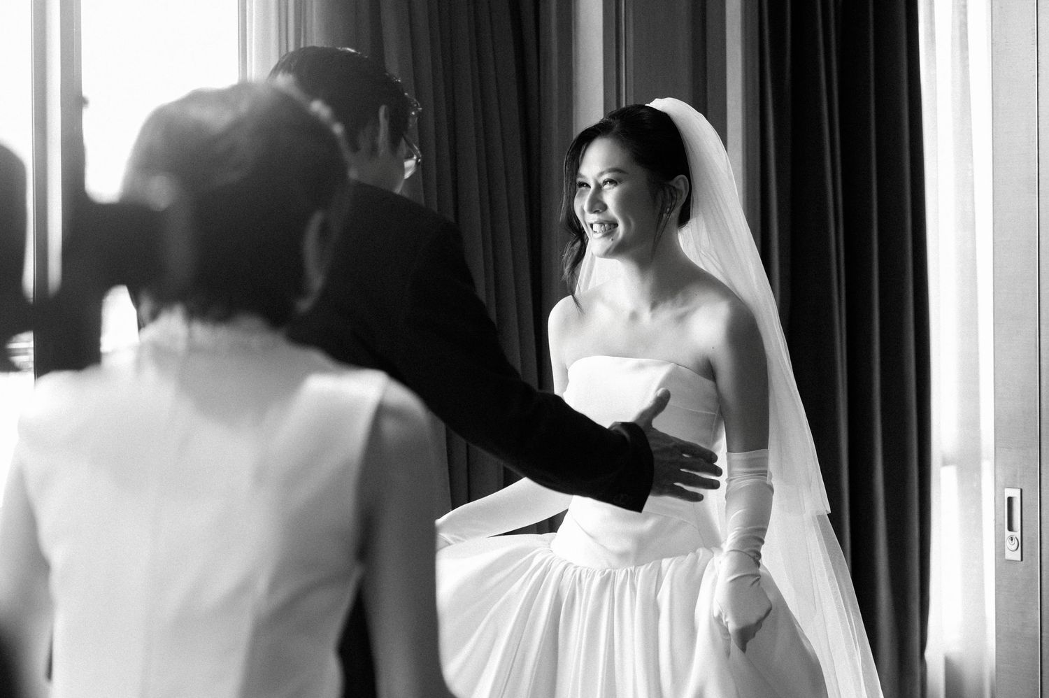 Black and white photo of a smiling bride in a strapless gown and veil during a wedding ceremony.