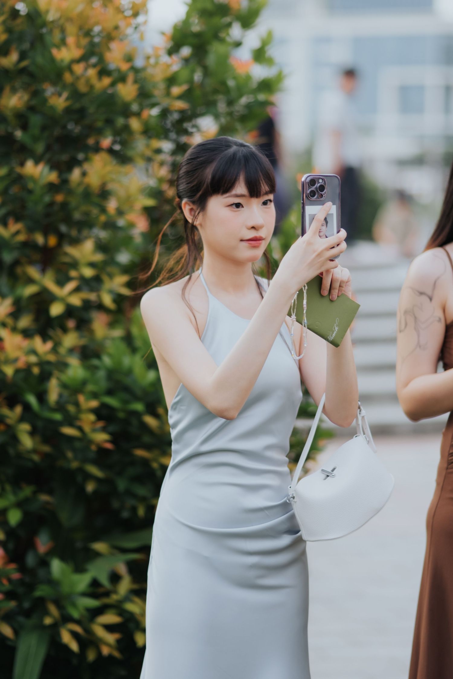 Young Asian woman in a light gray slip dress taking a photo with her smartphone outdoors near green bushes.