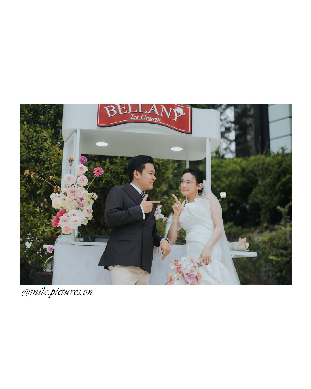 Couple in wedding attire sharing ice cream at a Bellany Ice Cream stand outdoors, surrounded by floral decorations.