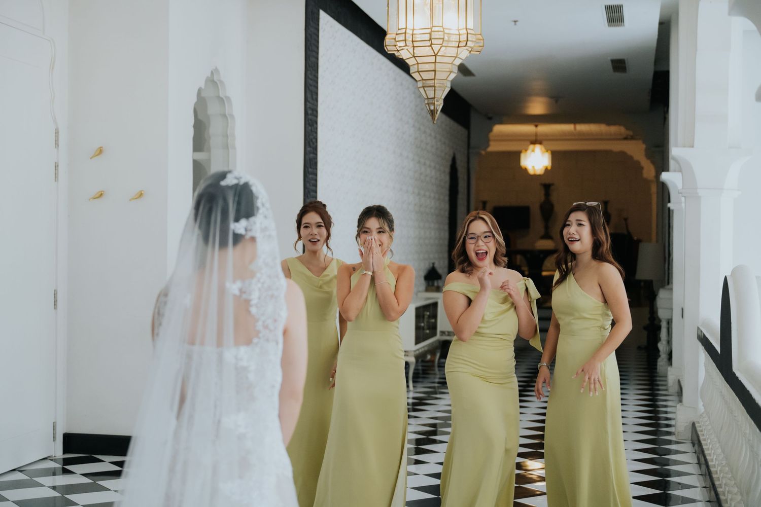 Bridesmaids in olive green dresses react with joy and surprise seeing the bride in her wedding gown in an elegant hallway.